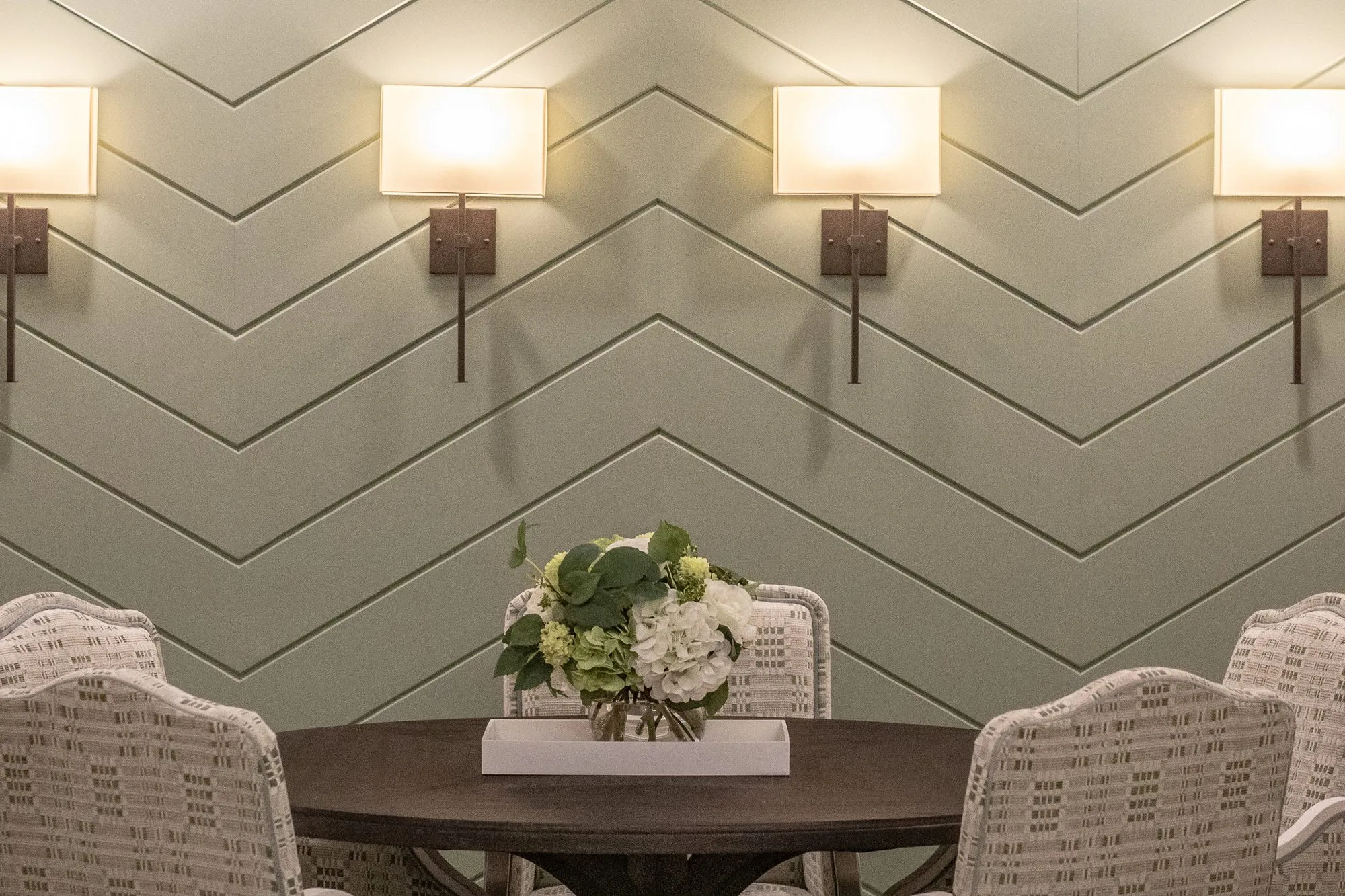 Senior living interior detail: round table with patterned skirted chairs and a floral centrepiece before a sage chevron wall