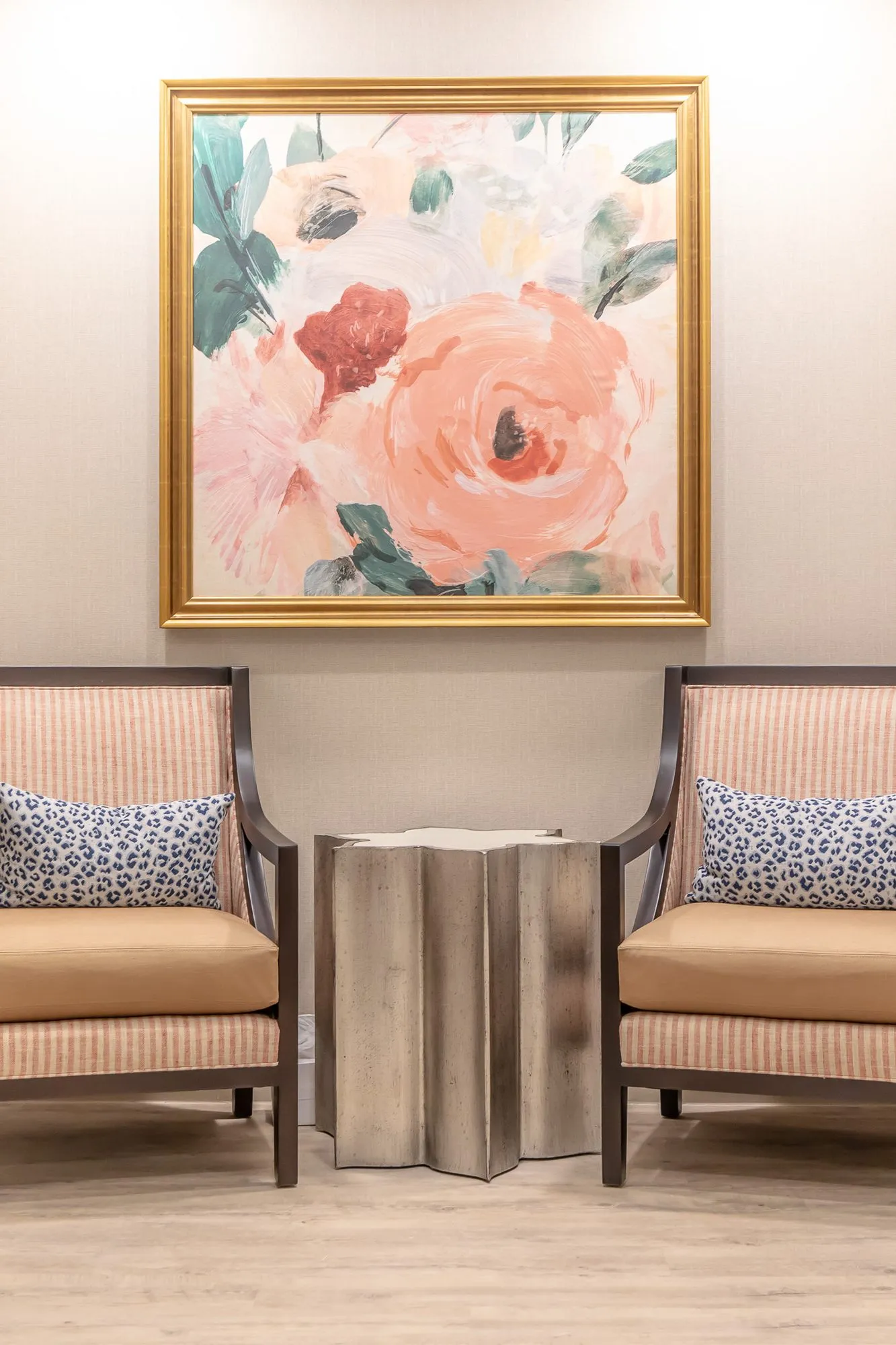 Senior living interior detail: striped armchairs with leopard print pillows and a silver side table below a floral painting
