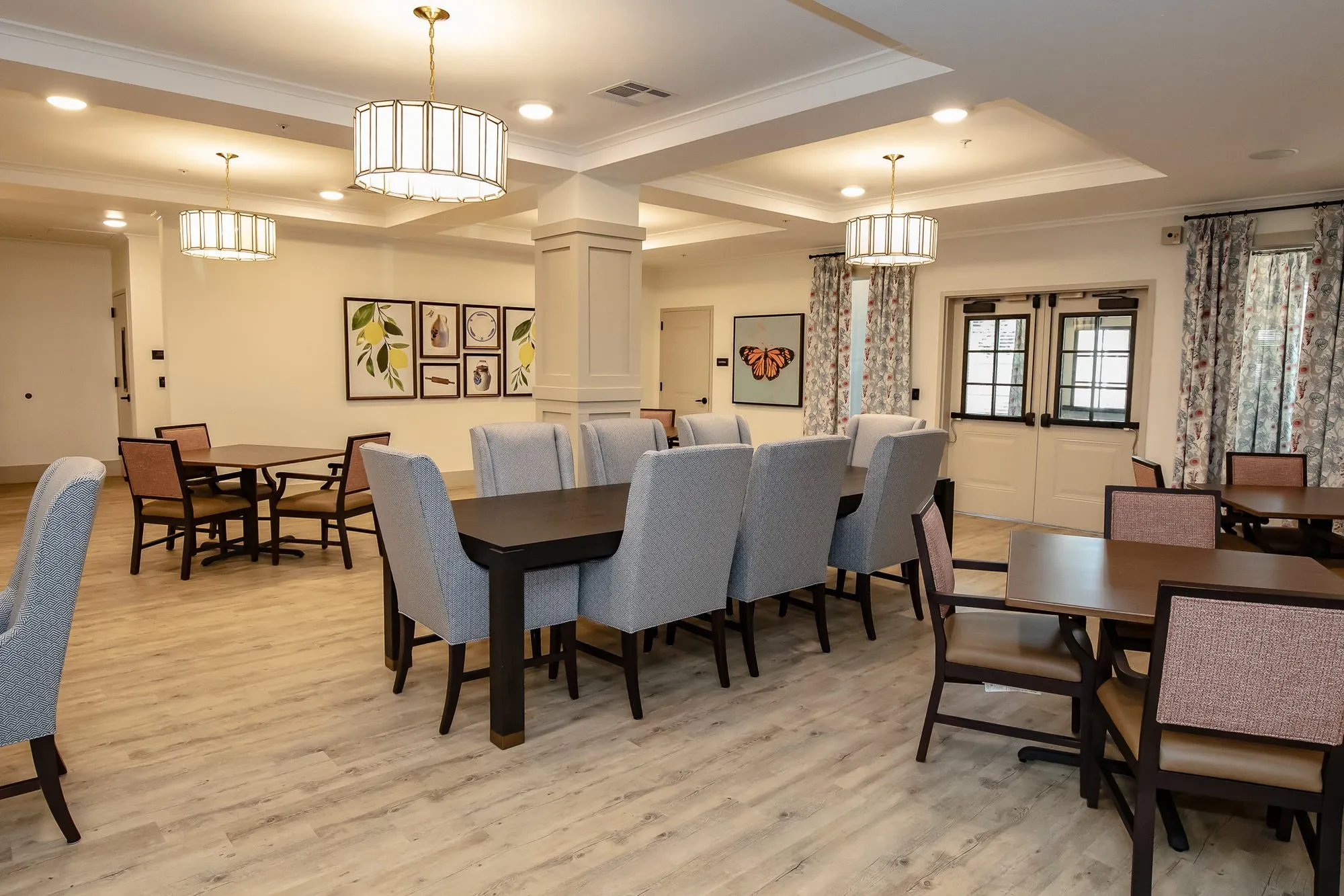 Senior living dining room toward a pair of French doors, a long table with grey chairs and botanical prints at left