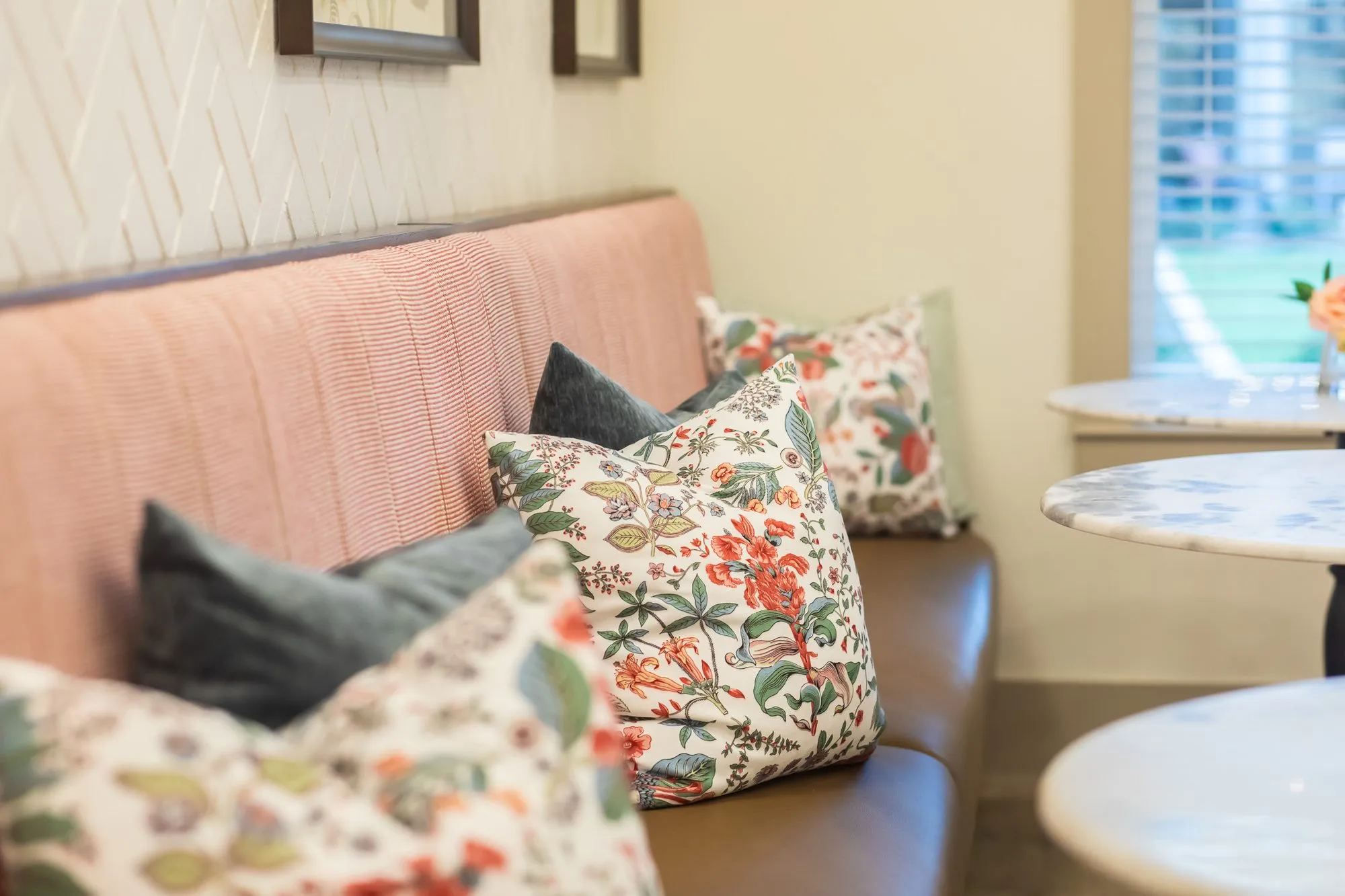 Senior living interior detail: floral and grey pillows on a pink channel tufted banquette beside marble bistro tables