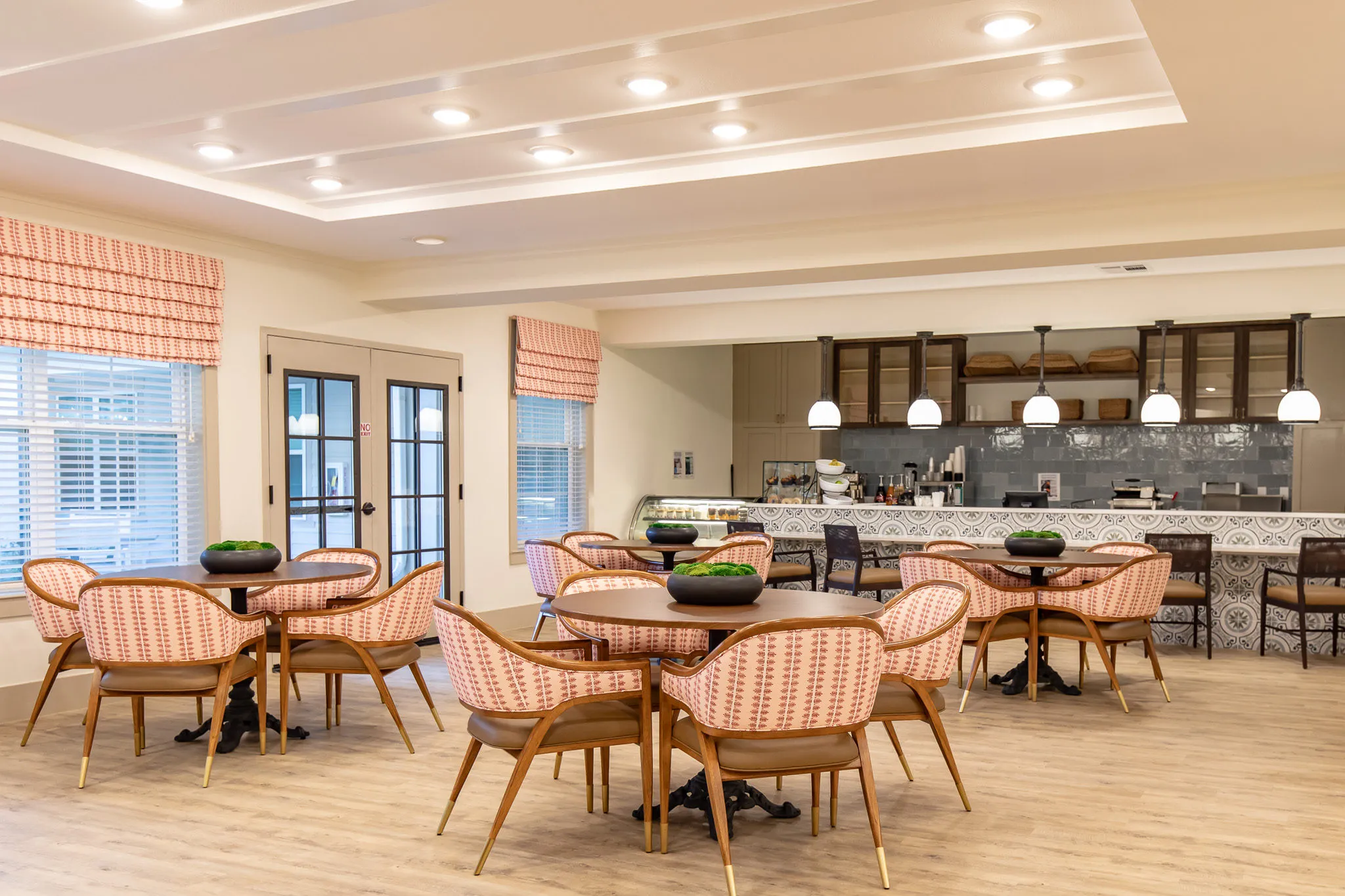 Senior living bistro with coral barrel chairs at round tables facing the open servery and patterned tile back bar