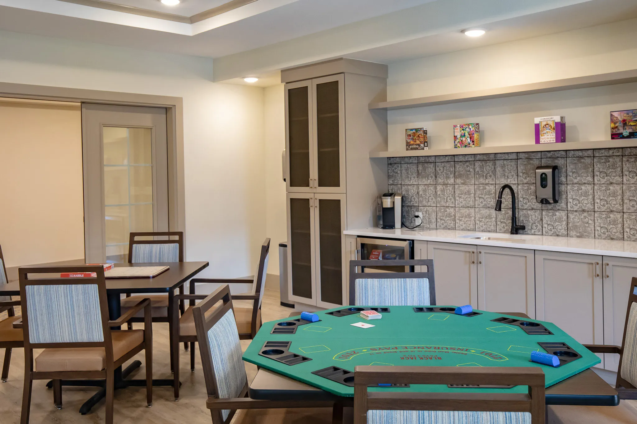 Senior living card room with a felt poker table and games tables beside the pantry counter and coffee station
