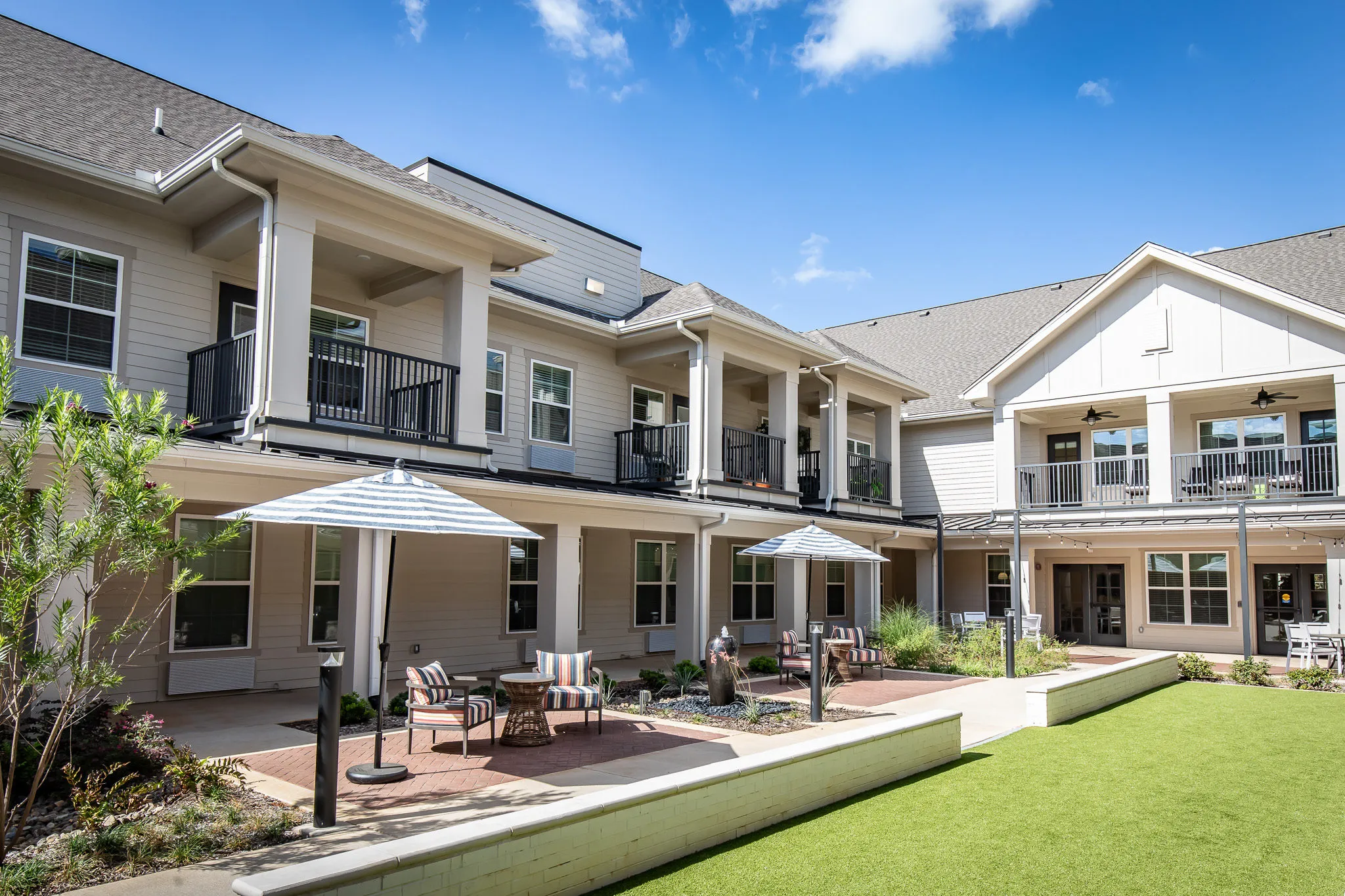 Senior living courtyard with market umbrellas and patio seating on brick paving beside the lawn and balconies
