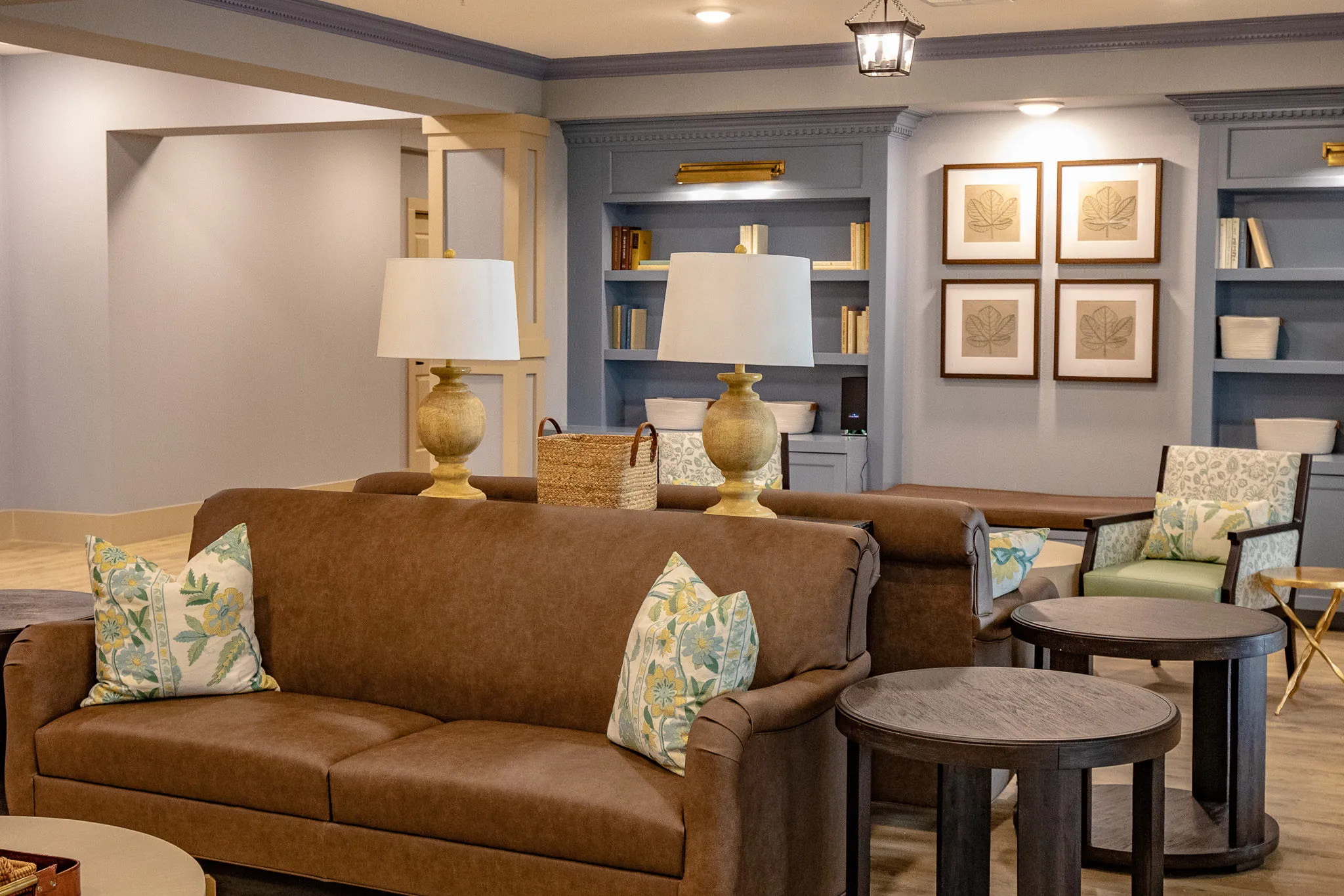 Senior living library lounge with a brown leather sofa and botanical pillows below blue-grey bookcases and framed prints