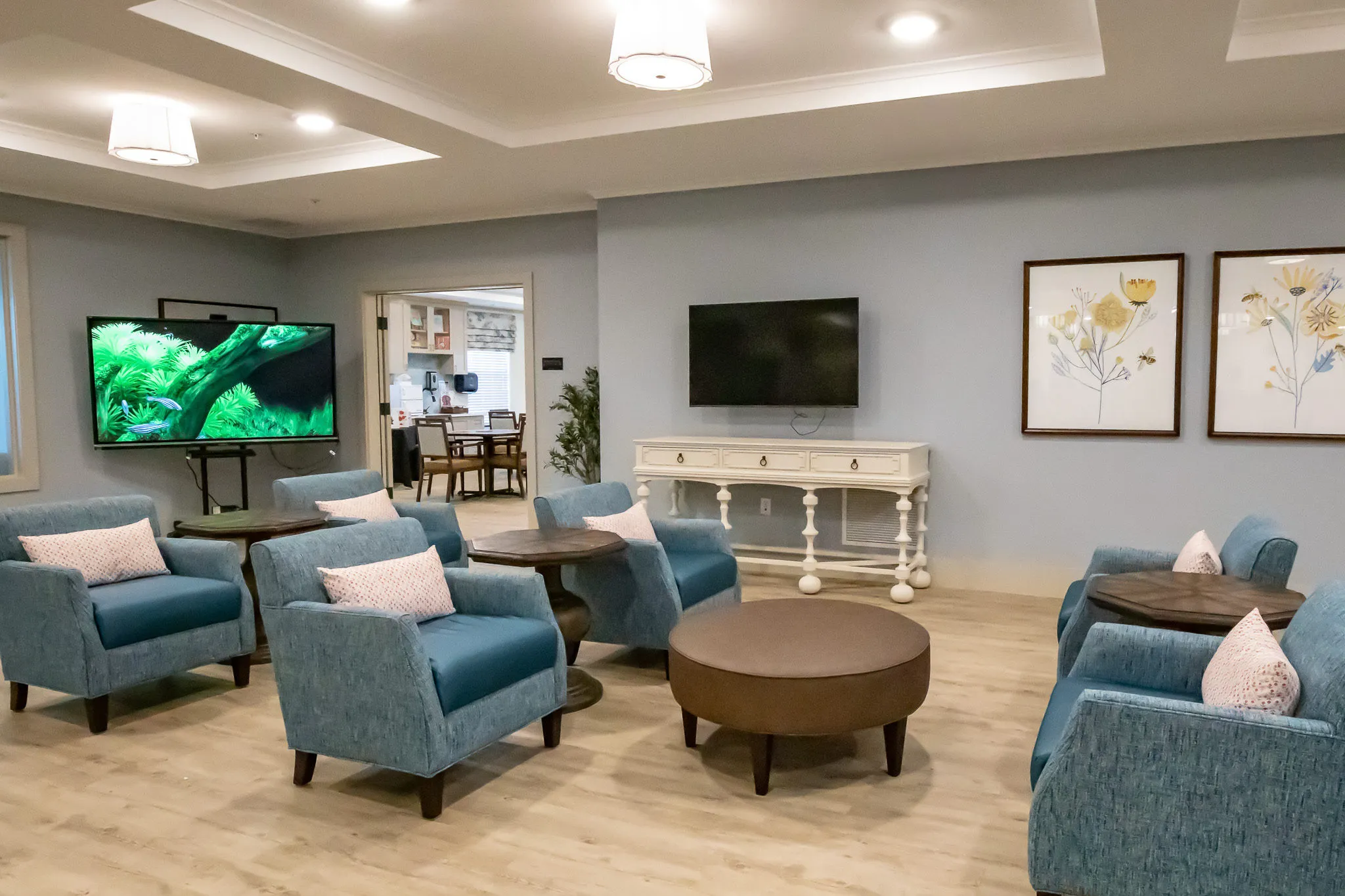 Senior living lounge with teal armchairs around round wood tables below a TV wall, a white console alongside