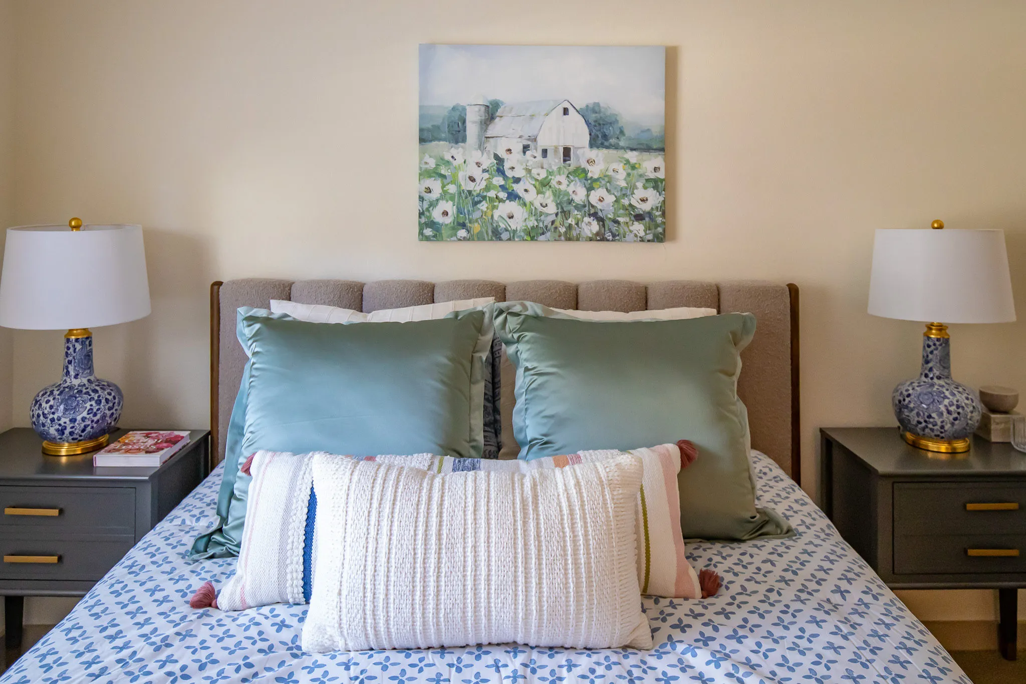 Senior living model bedroom with teal shams and a blue floral coverlet on the upholstered bed below a barn painting