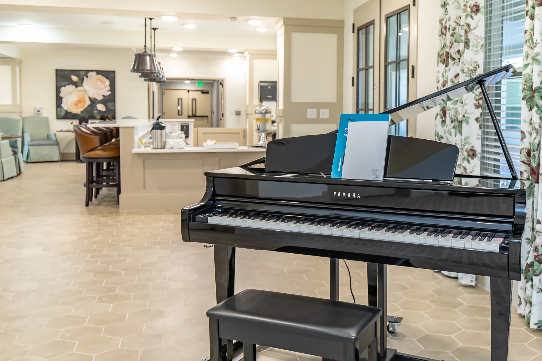 Senior living lounge with a grand piano on hexagonal tile beside floral drapery, a bistro counter and armchairs beyond
