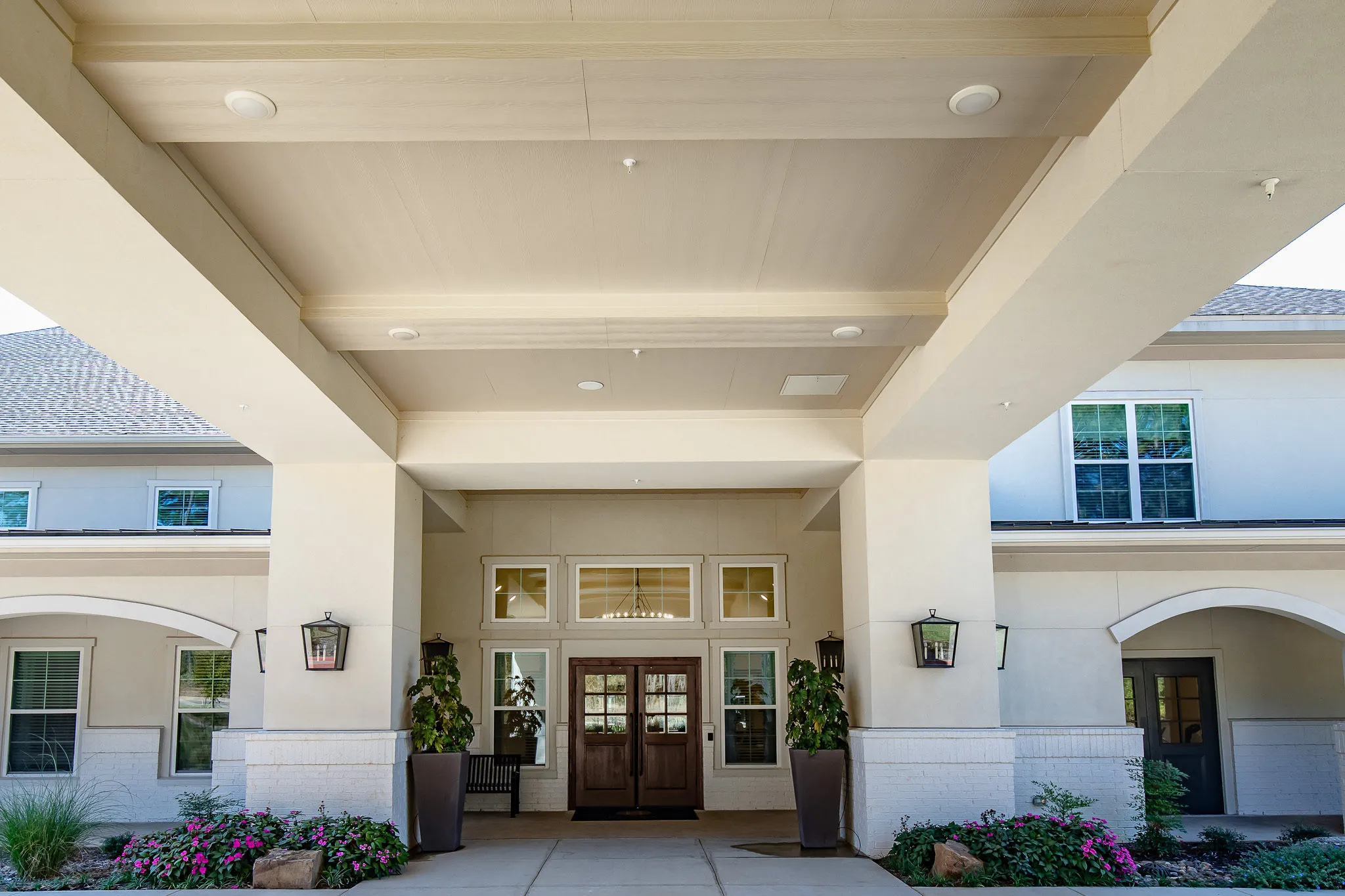 Senior living community exterior: arrival court with timber double doors and lanterns under the covered porte cochere