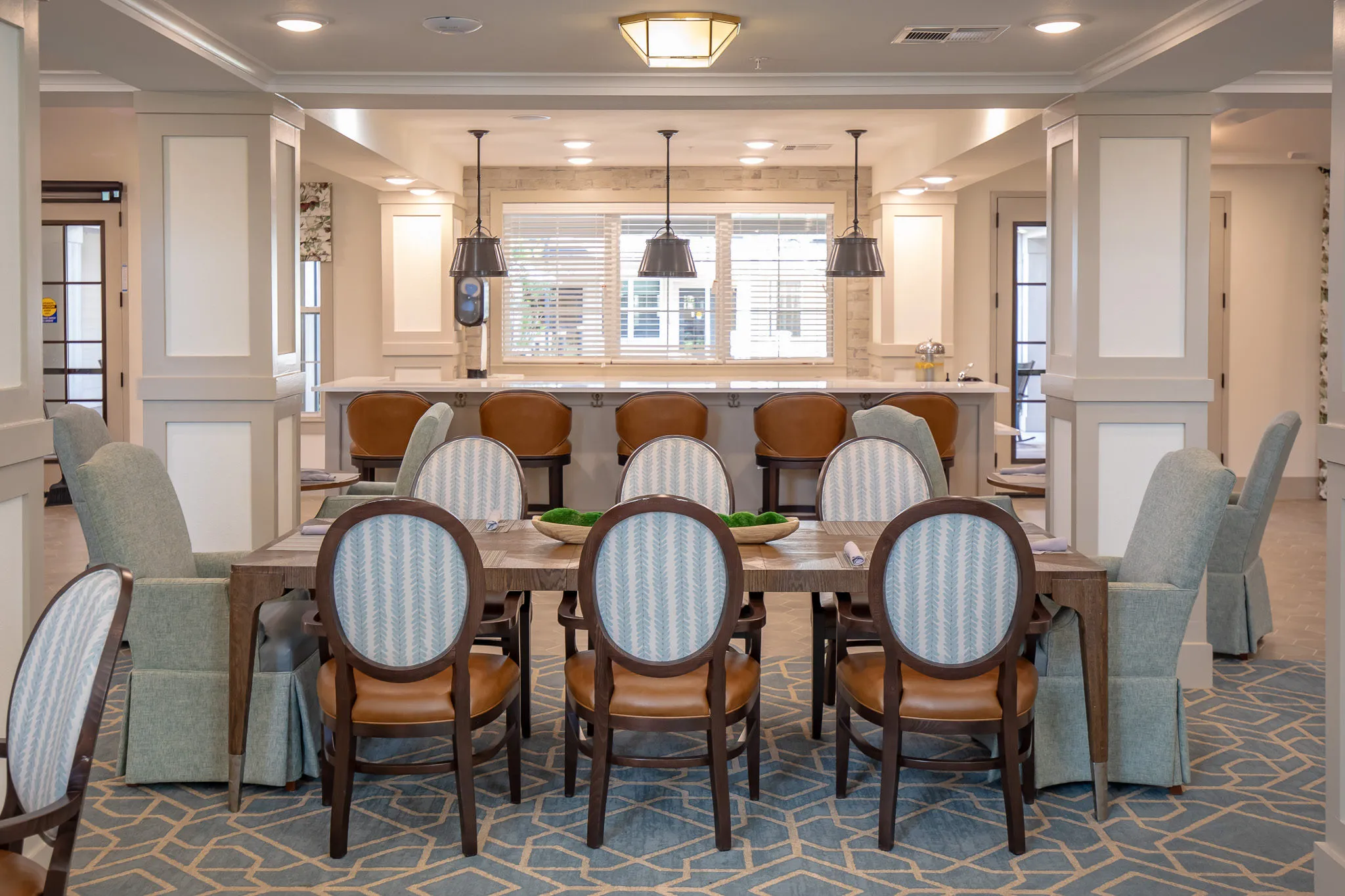 Senior living dining room with oval-back and skirted chairs along a plank table, an island under black dome pendants beyond