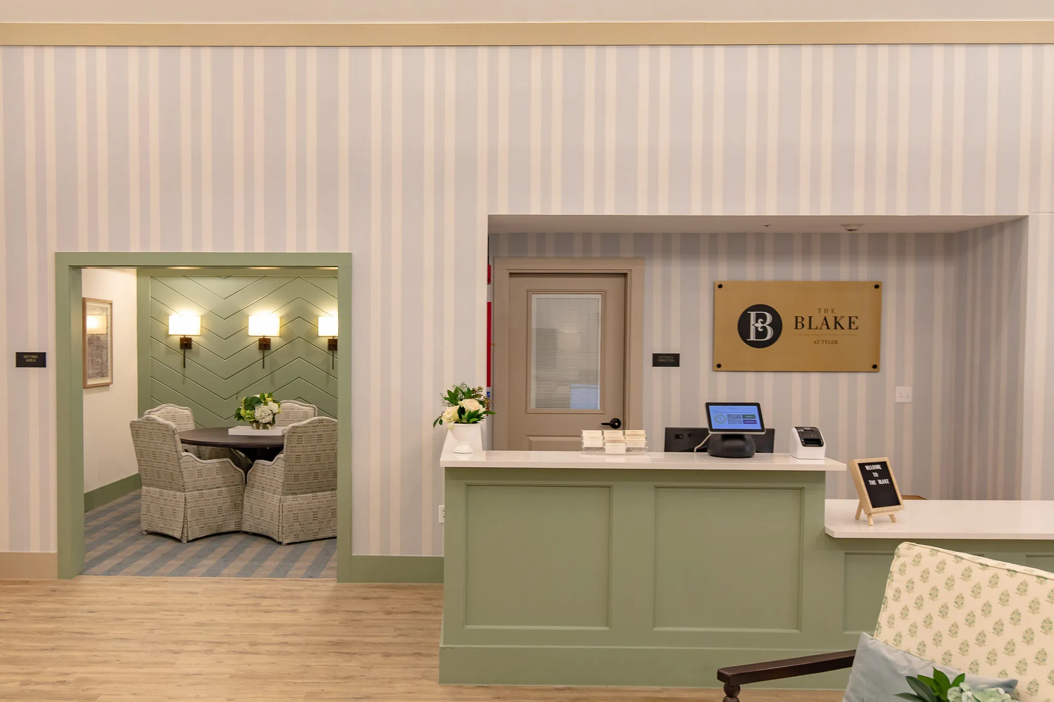 Senior living reception with a sage green desk against striped wallcovering, the chevron-panelled sitting room beyond