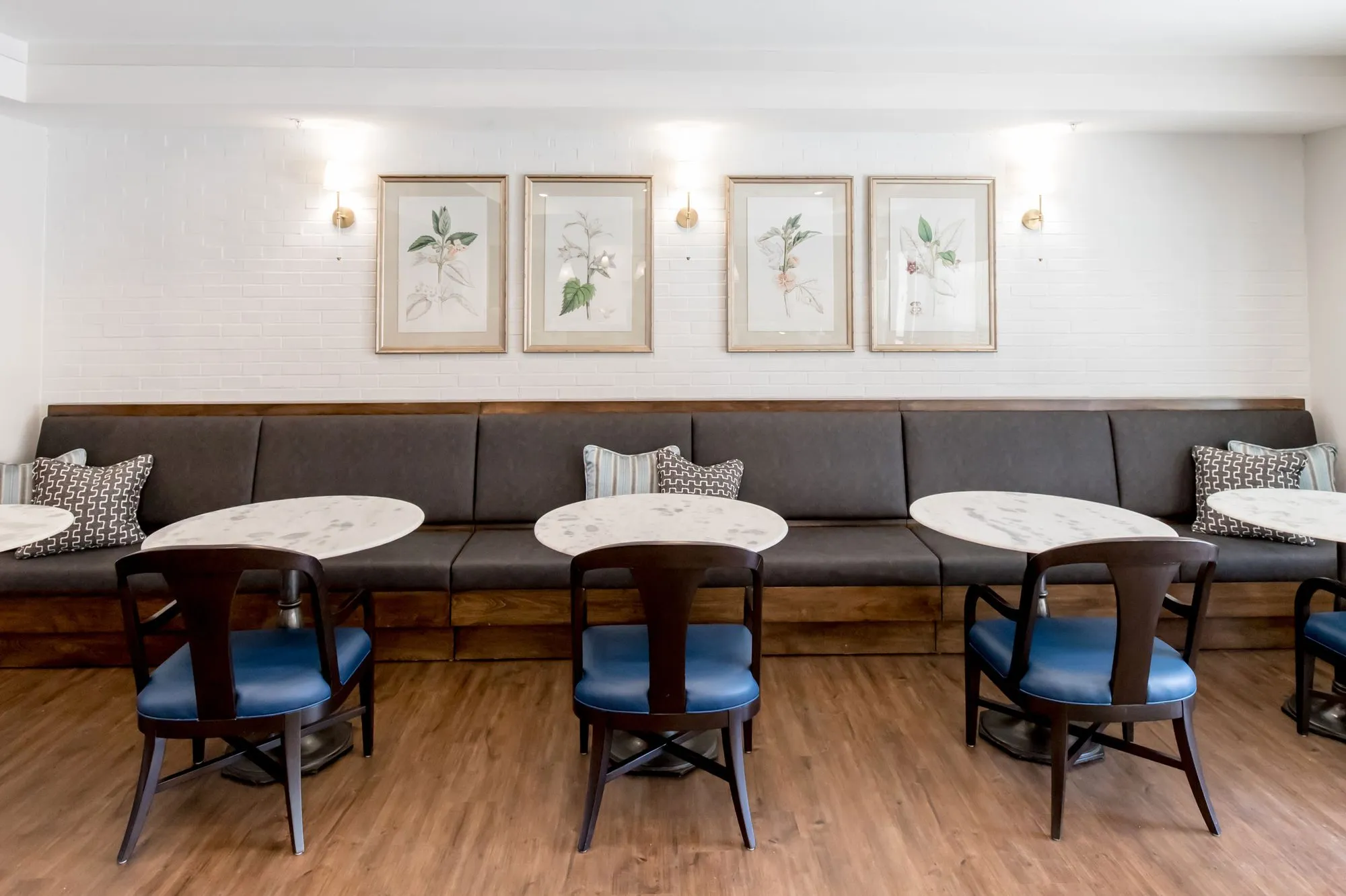 Senior living dining area with a long charcoal banquette, marble tables and blue-seated wood chairs below botanical prints
