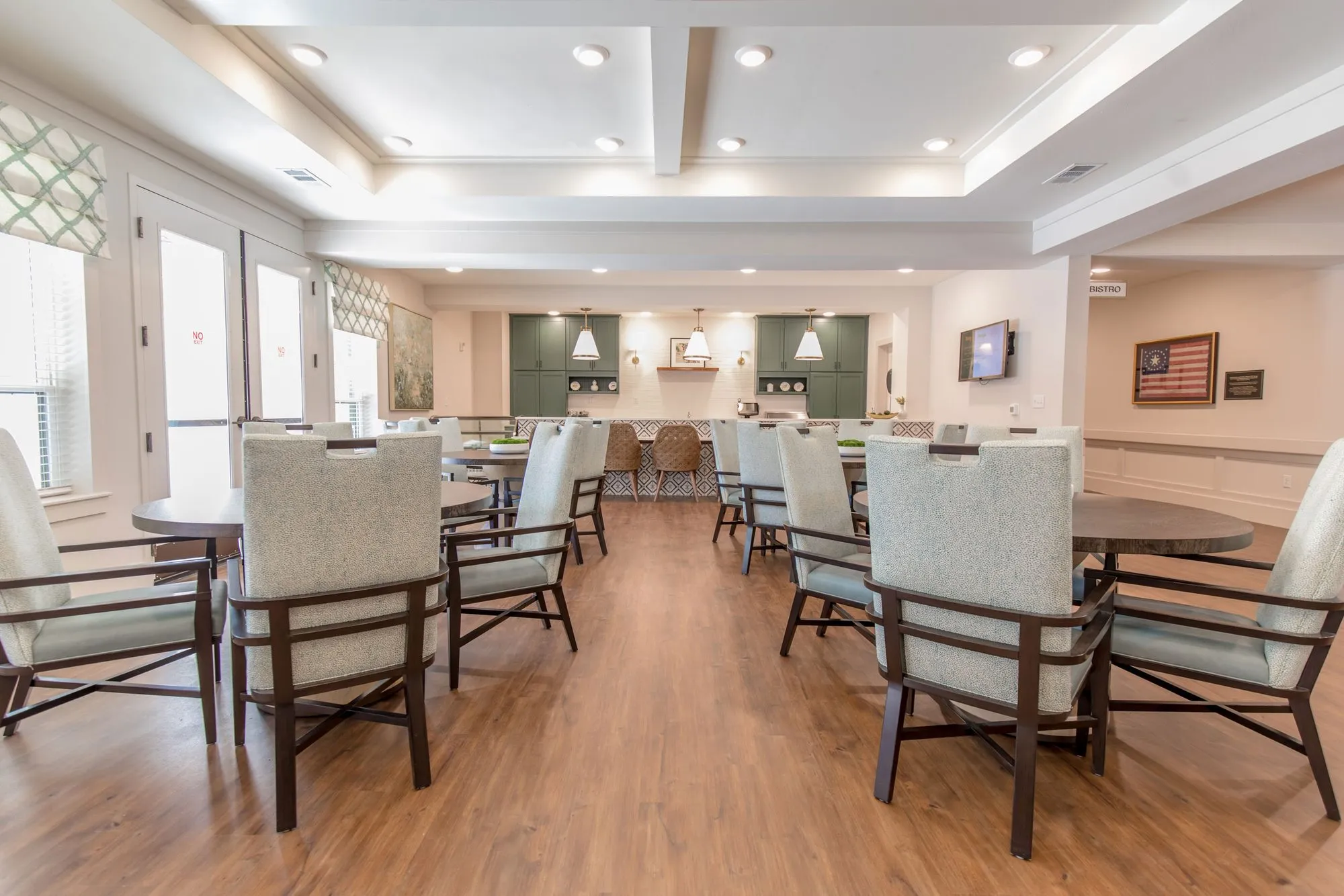 Senior living dining room with grey upholstered chairs at round tables under a coffered ceiling, sage cabinetry beyond