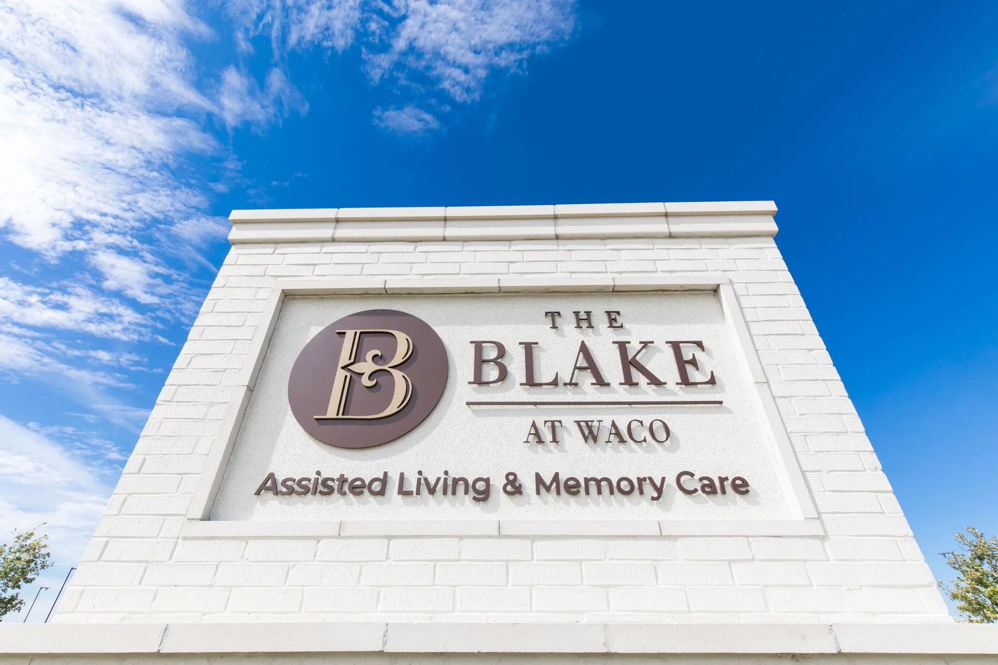 Senior living community exterior: white brick monument sign with a round monogram logo against a blue sky