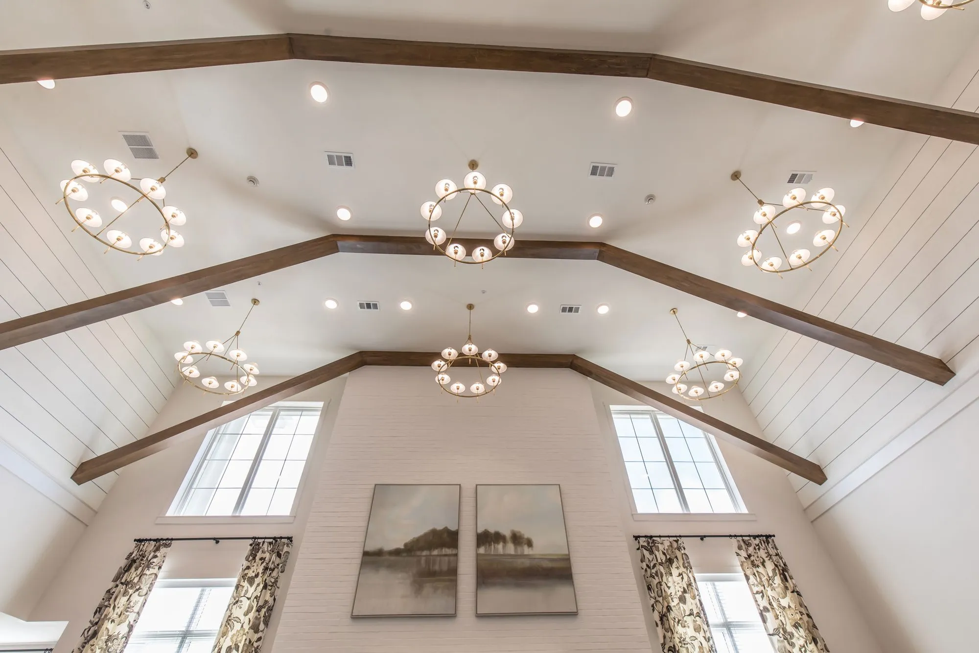 Senior living interior detail: vaulted plank ceiling with timber beams and ring chandeliers above tall windows and paintings