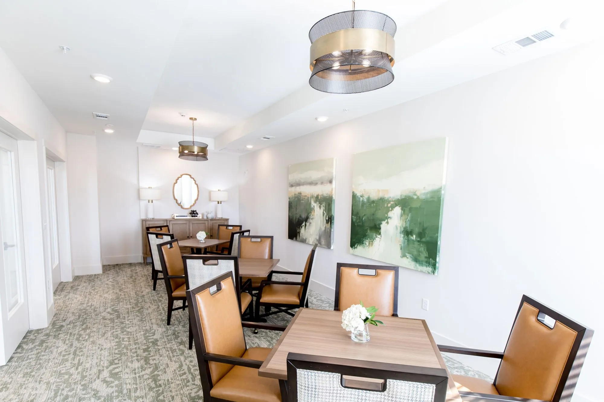 Senior living private dining room with cognac leather chairs at wood tables, drum pendants and two green landscape paintings