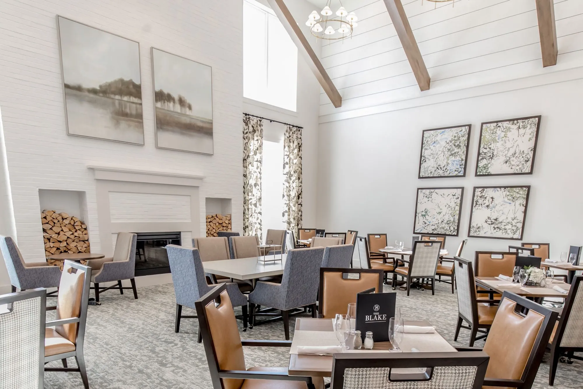 Senior living dining room angled toward the fireplace and stacked firewood, cognac and grey chairs at set tables