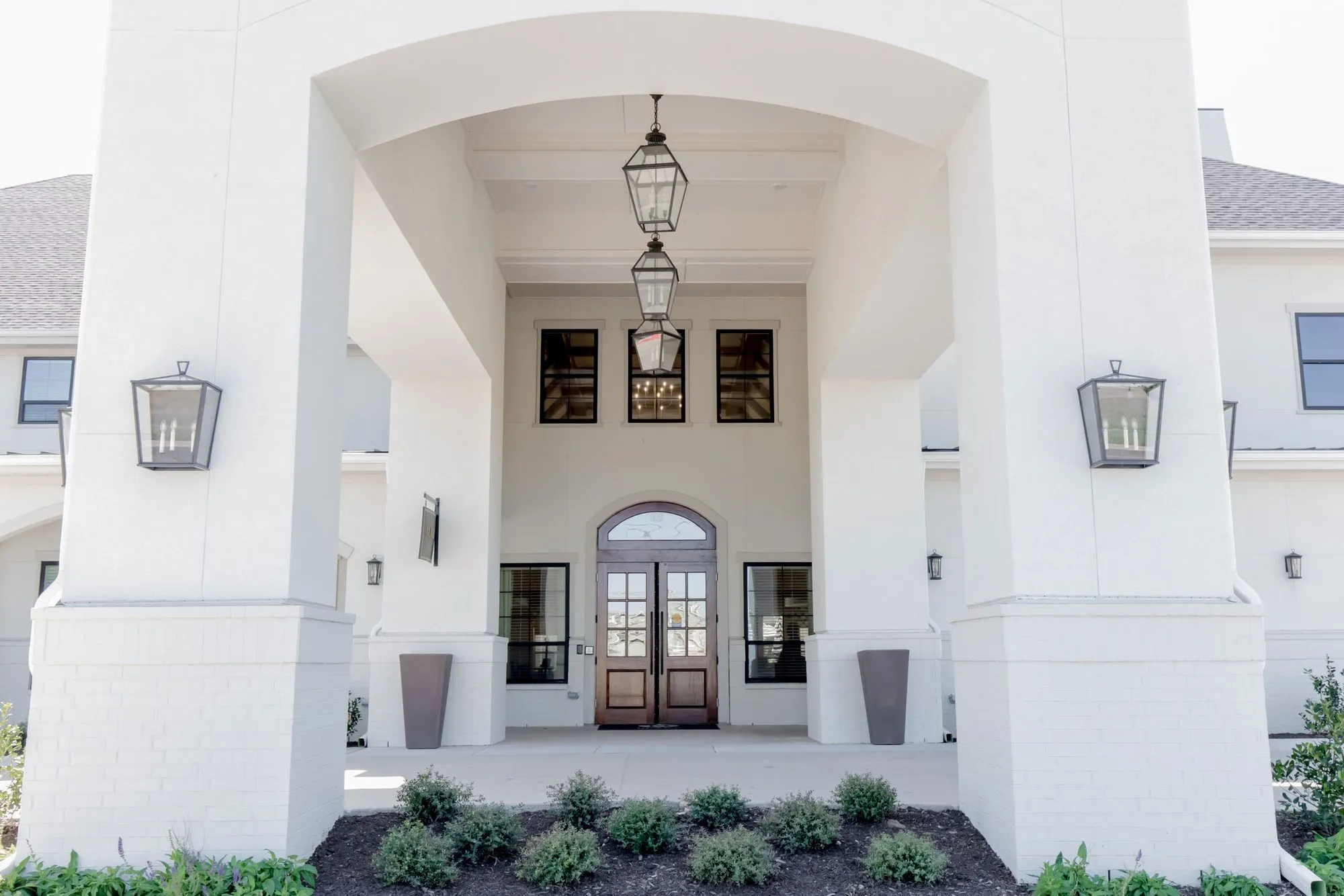 Senior living community exterior under the arched white porte cochere, lanterns above the arched double entry doors