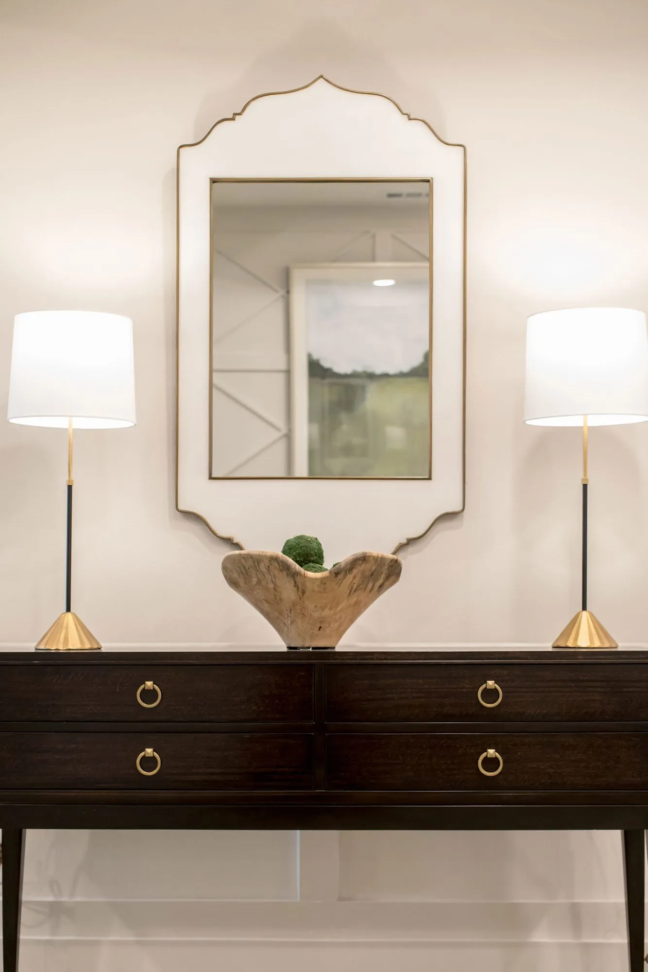 Senior living interior detail: dark console with a wood bowl between two white lamps below a scalloped white mirror