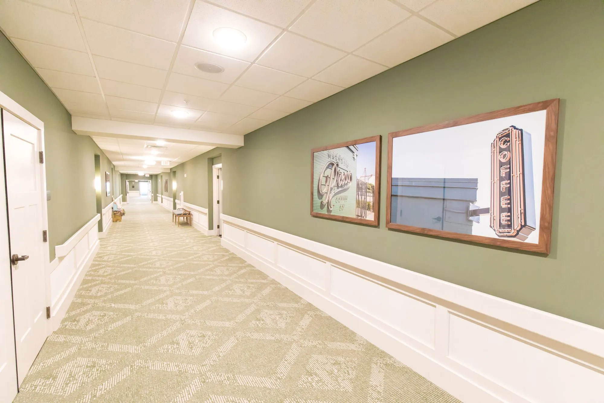 Senior living corridor in sage with white wainscot and patterned carpet, framed photographs of vintage signs along the wall