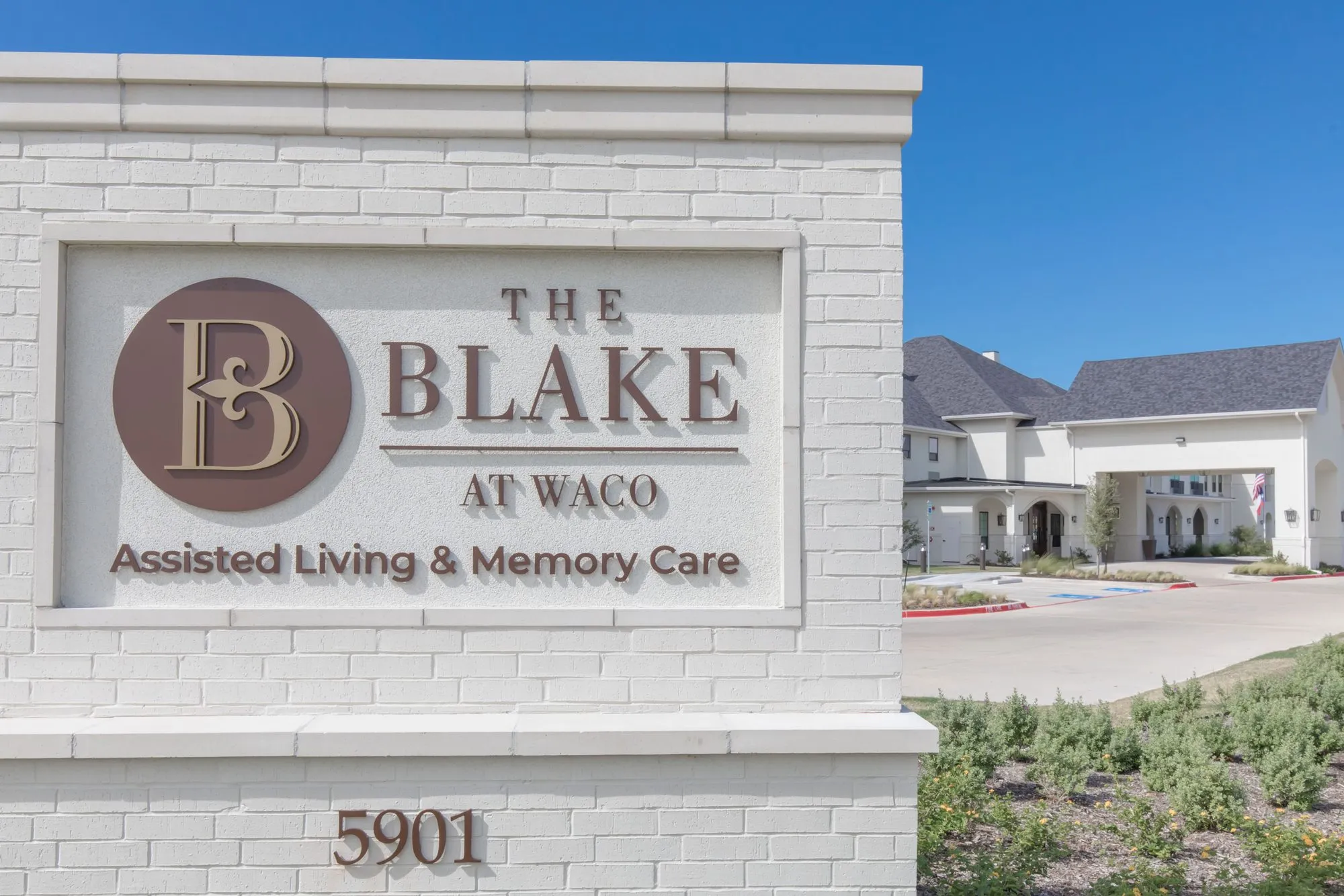 Senior living community exterior: white brick monument sign by the drive, the two storey building and porte cochere beyond