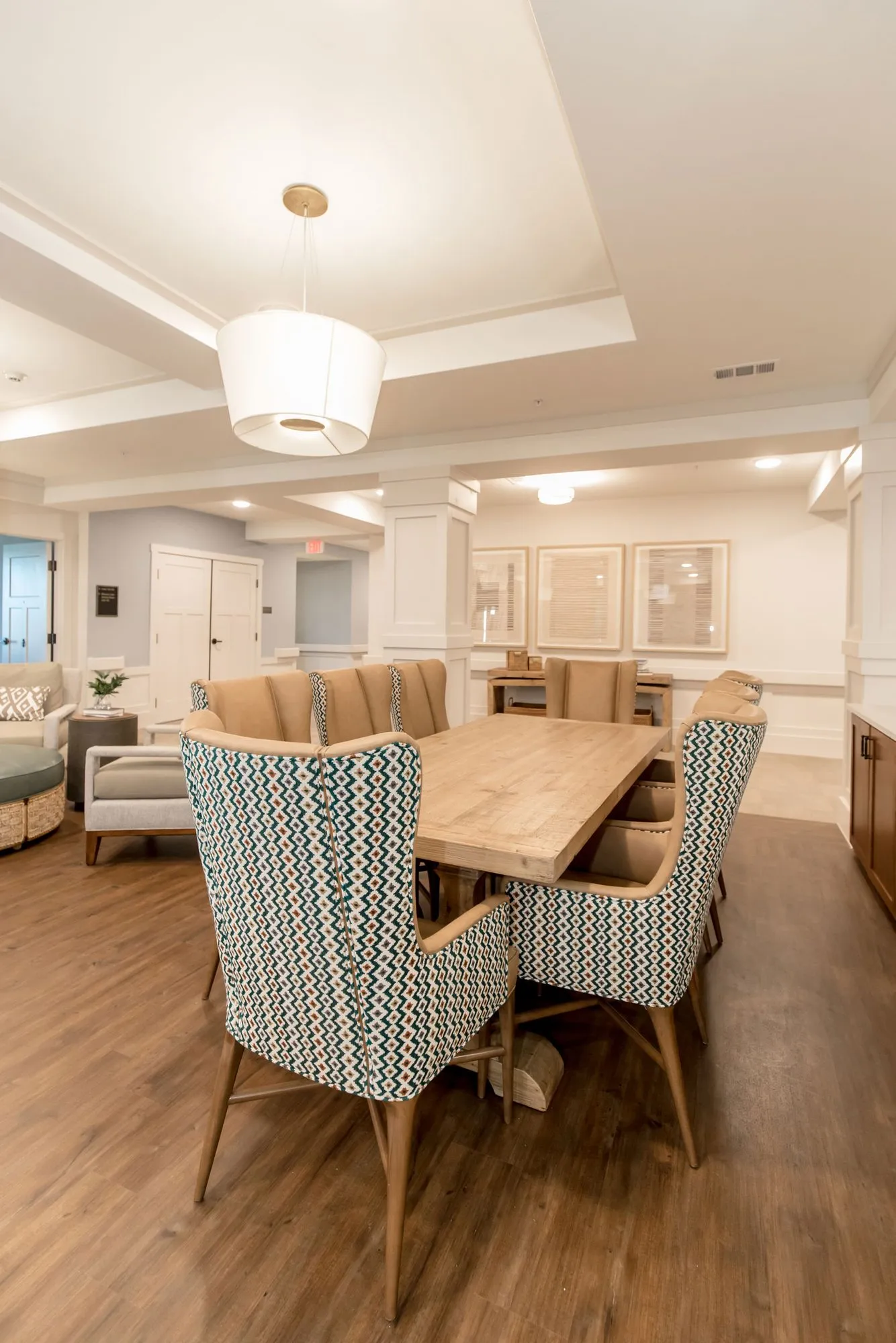 Senior living dining area with blue patterned wing chairs at a long wood table under a drum pendant, the lounge beyond