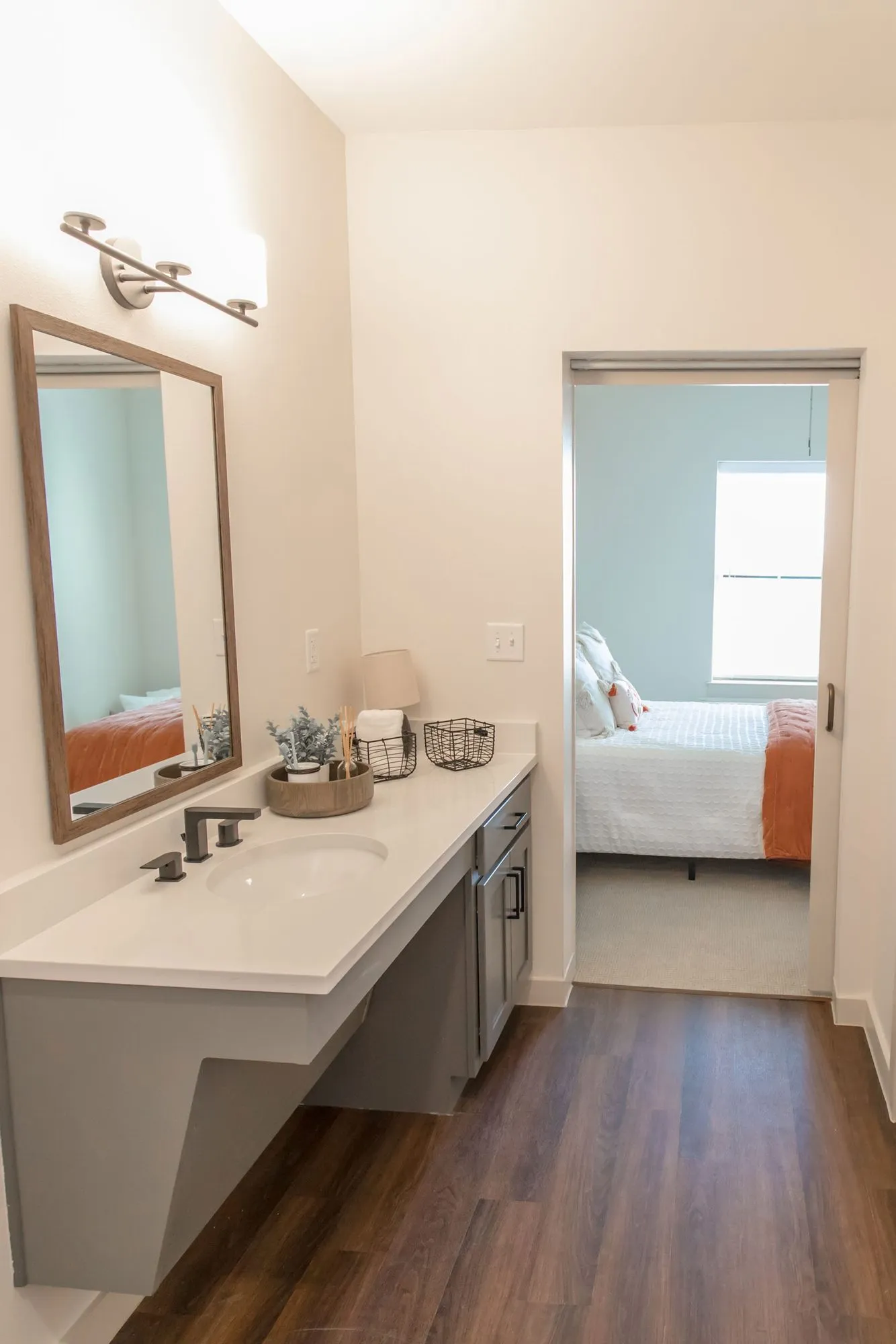 Senior living apartment bathroom with an accessible vanity, wood-framed mirror and bar light, the bedroom beyond