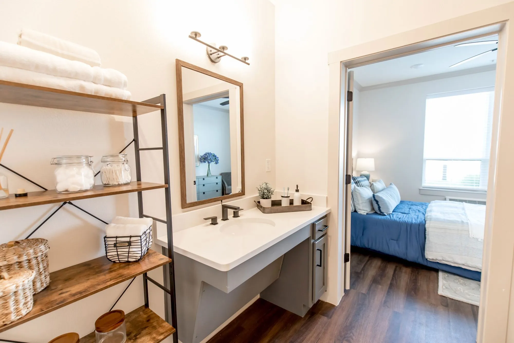 Senior living apartment bathroom with open shelving of towels beside an accessible vanity, a bedroom with blue bedding beyond