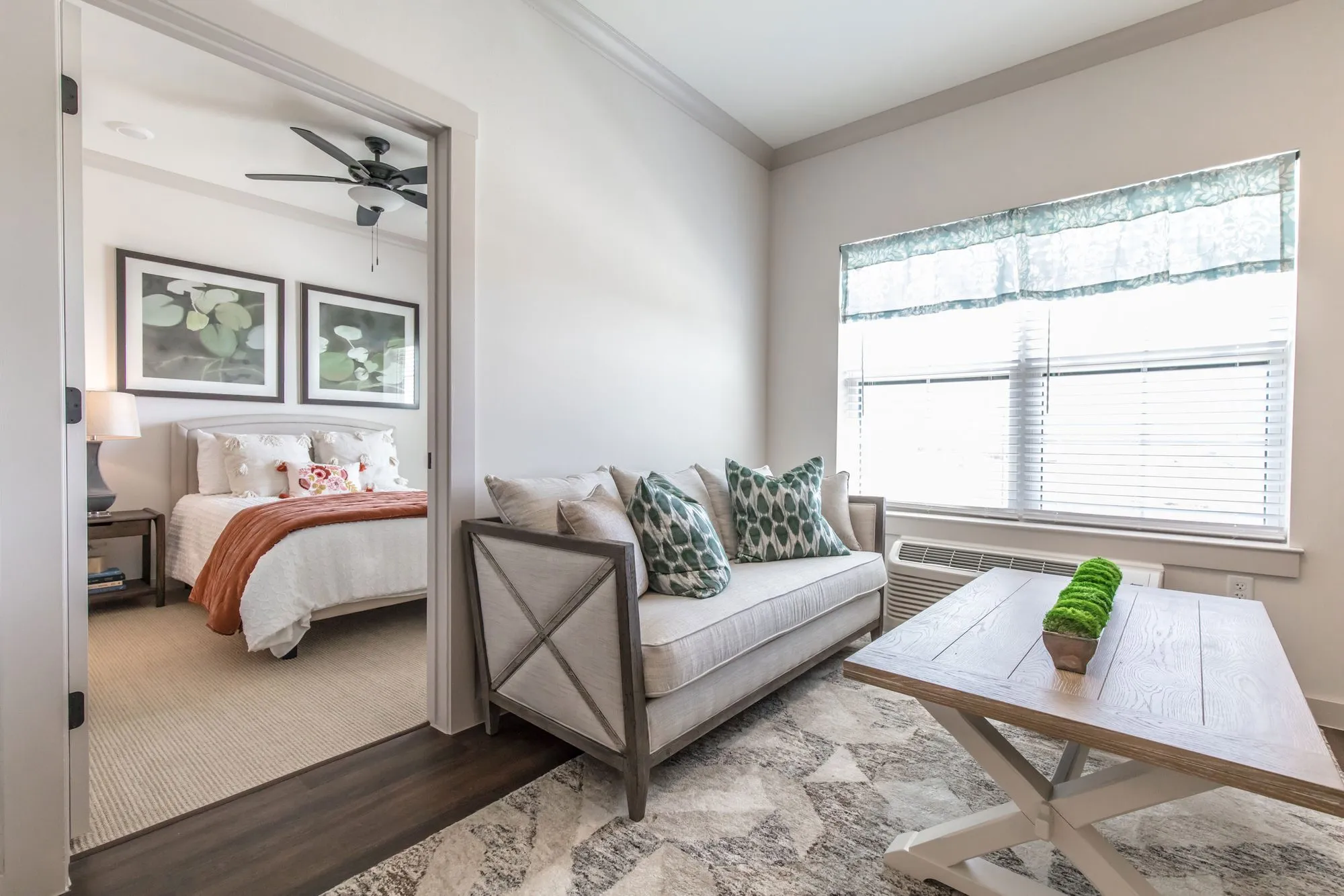 Senior living apartment living room with a linen sofa, green leaf pillows and a farmhouse coffee table, the bedroom beyond