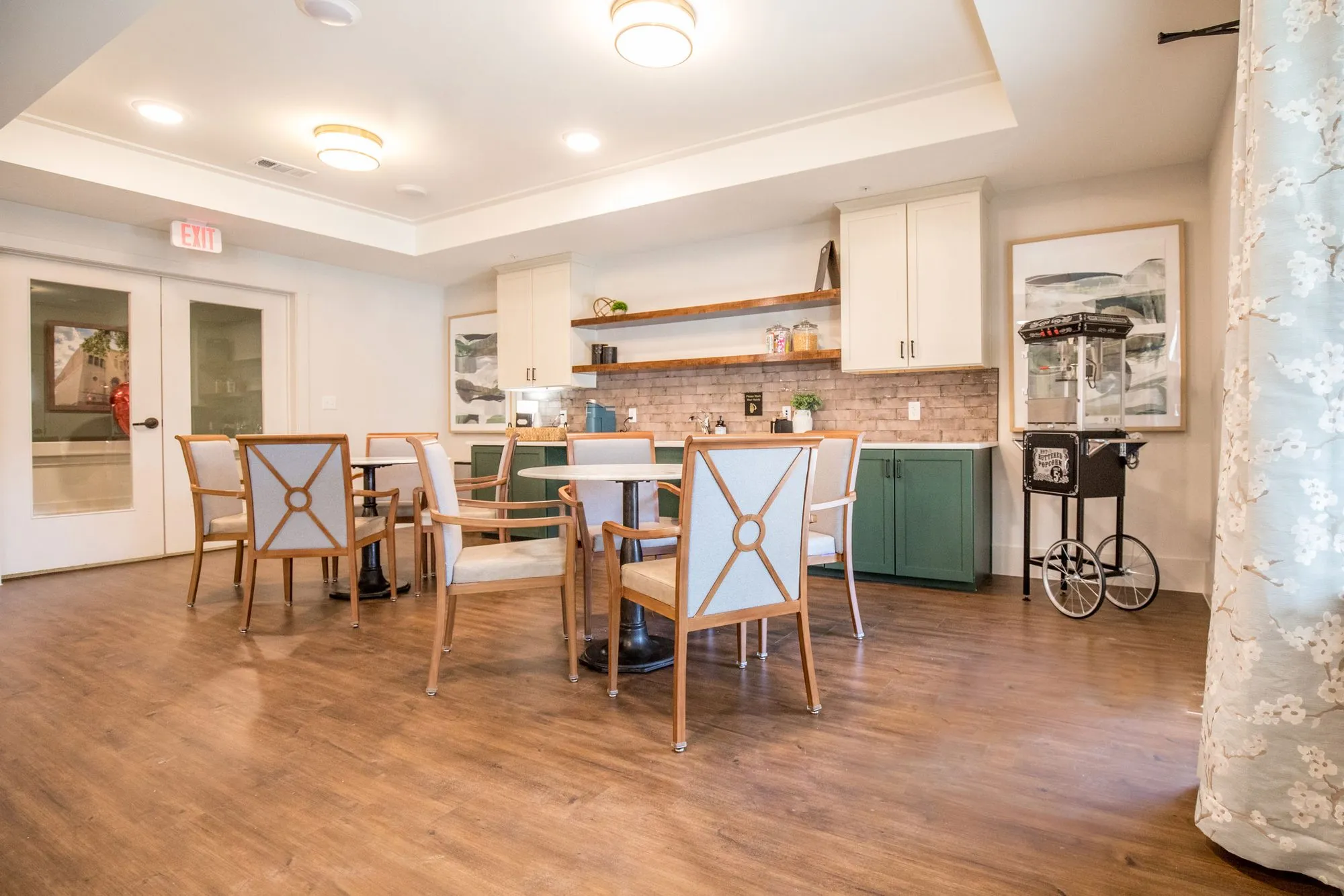 Senior living activity kitchen with wood tables and cream patterned chairs, green cabinetry, open shelves and a popcorn cart