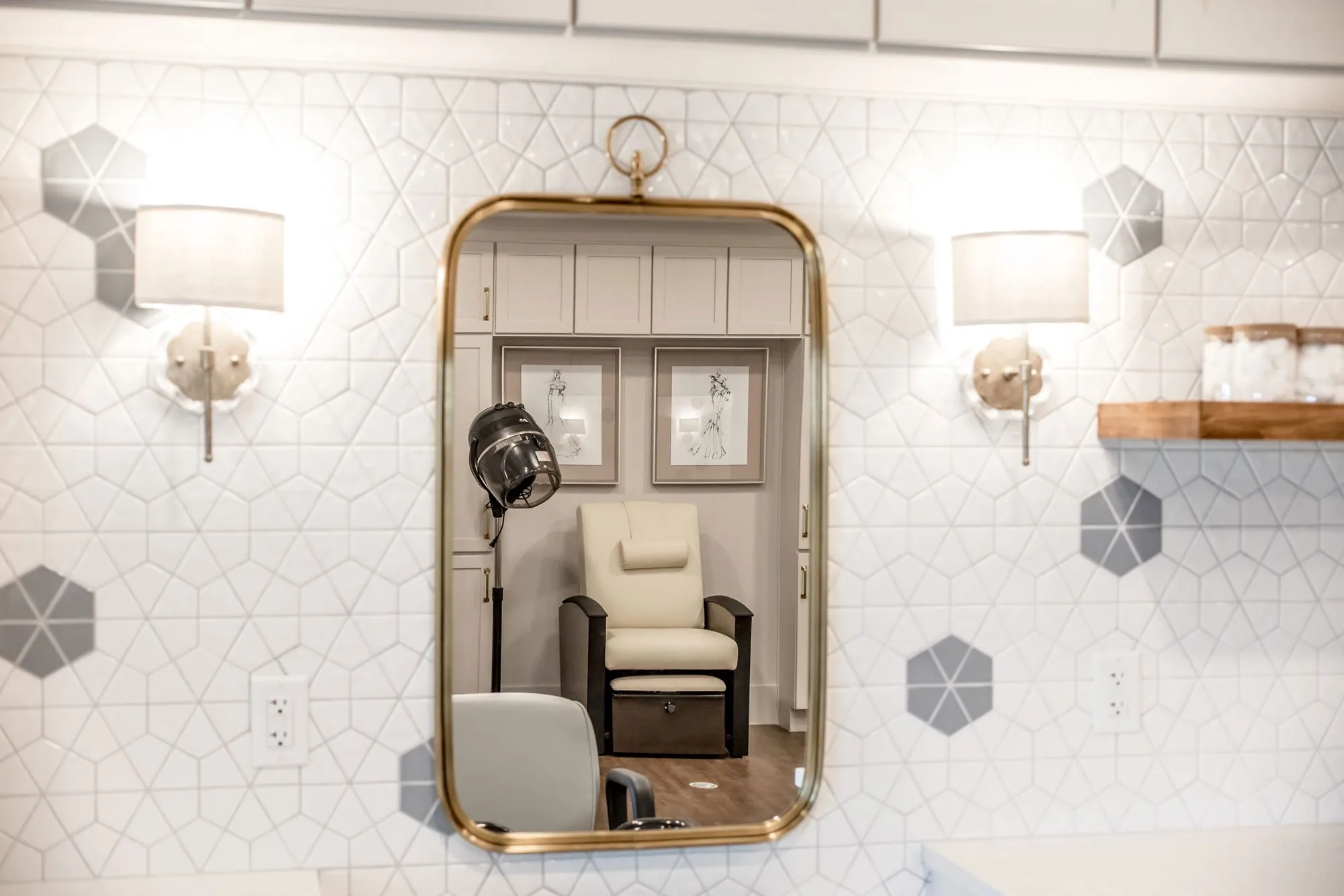 Senior living salon with a brass-framed mirror and linen sconces on white geometric tile, reflecting the pedicure chair