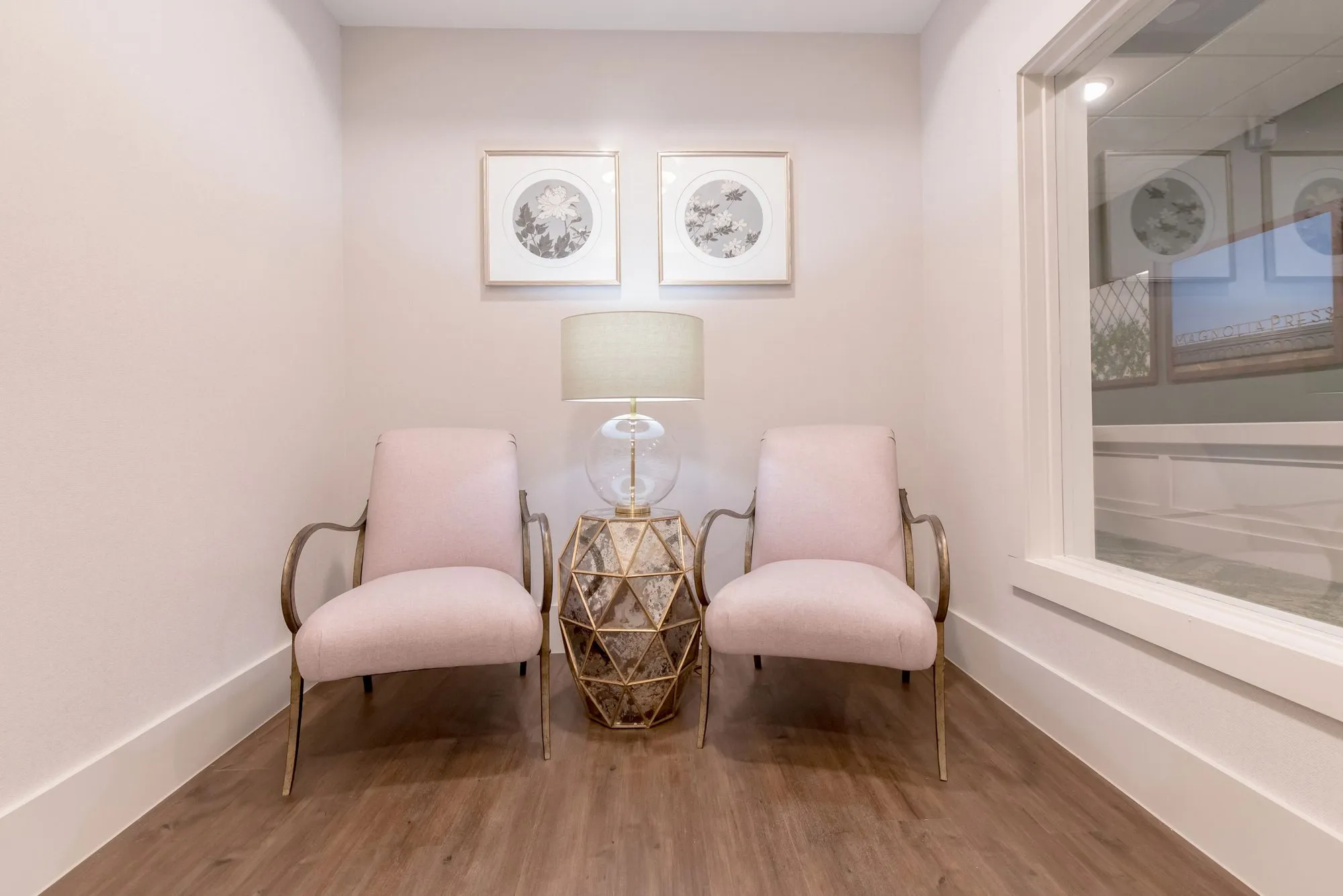 Senior living interior detail: pair of blush armchairs and a gold faceted side table with a glass lamp under botanical prints