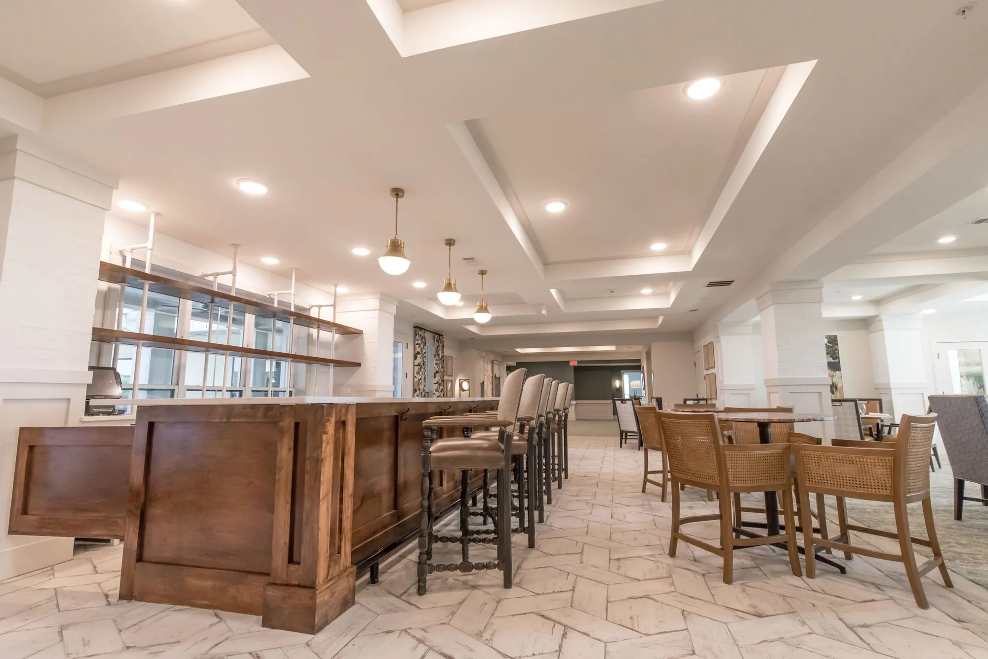 Senior living bistro with a wood-panelled counter and stools under pendants, woven chairs at tables on a herringbone floor