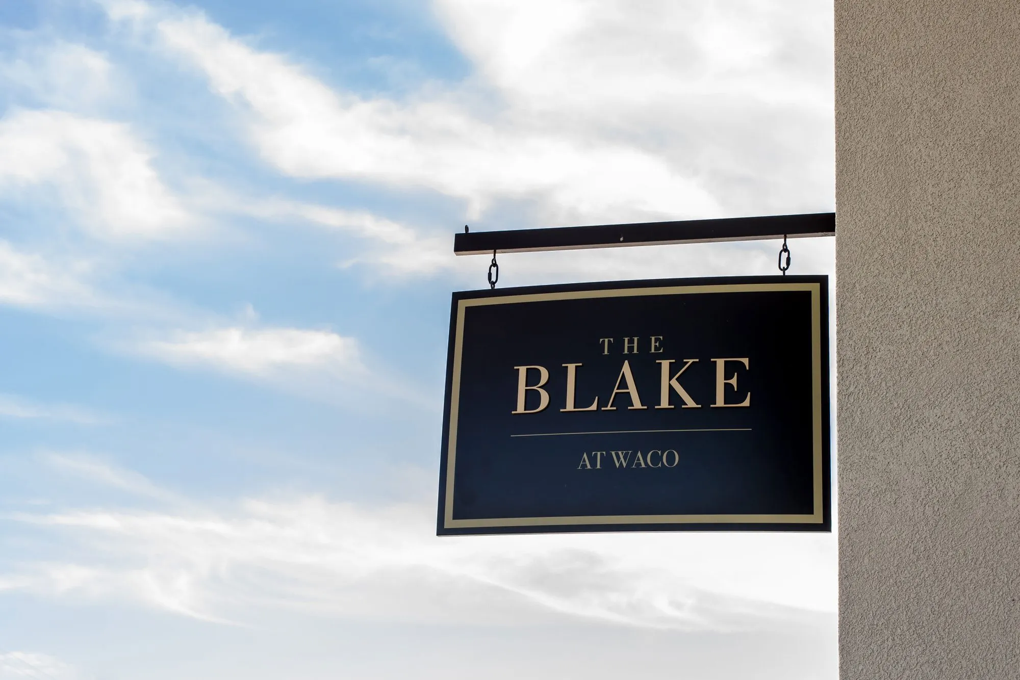 Senior living community exterior: black hanging blade sign with gold lettering on an iron bracket against a cloudy sky