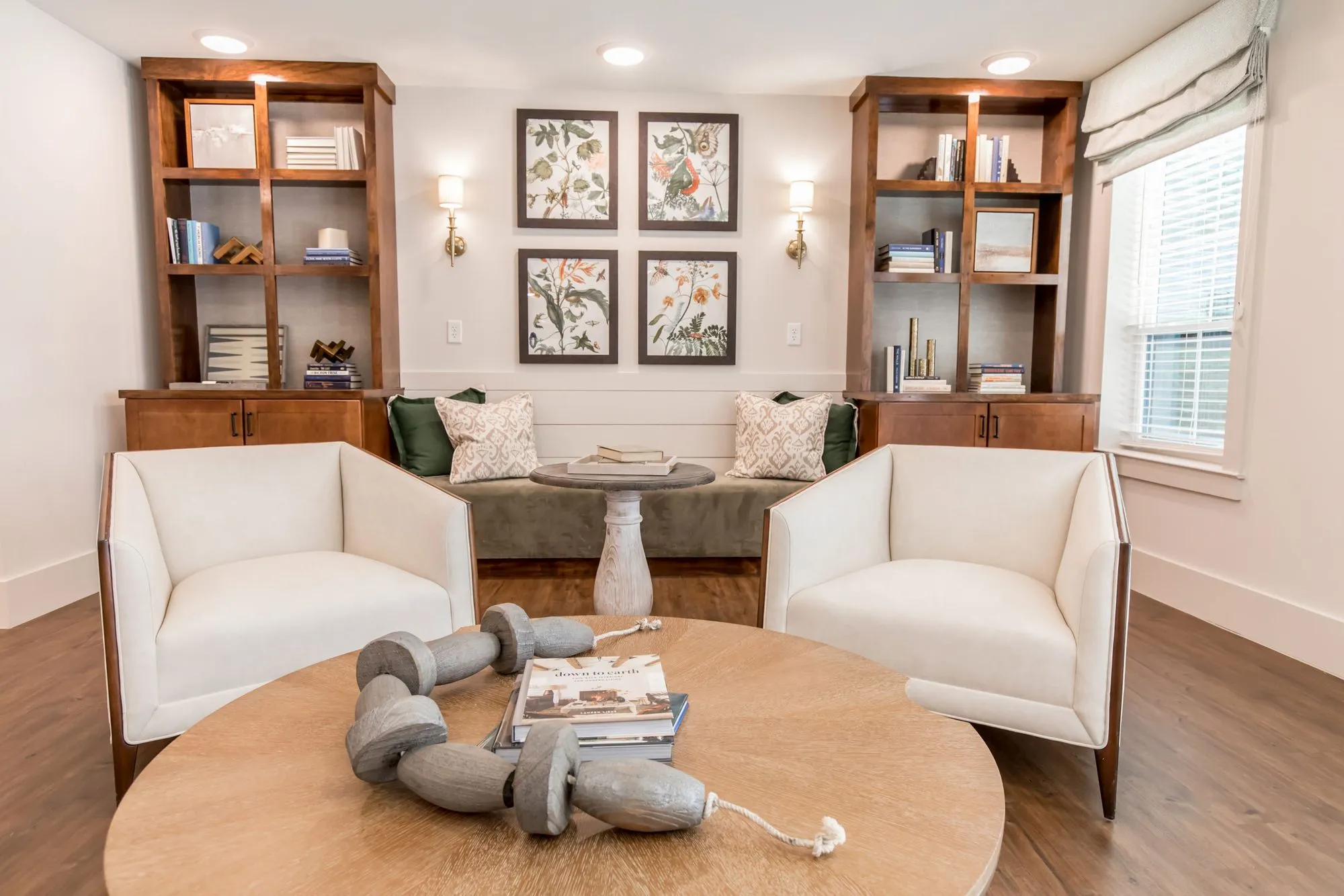 Senior living library nook with a round wood table between two white armchairs, the green banquette and prints beyond