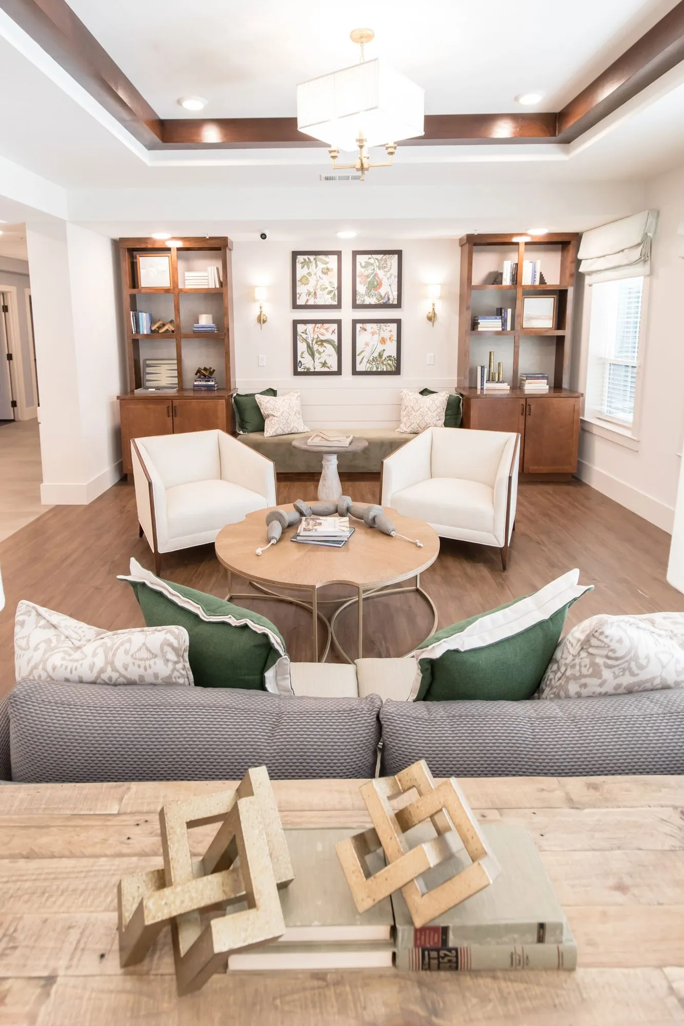 Senior living living room seen over a wood table with a fretwork sculpture, past a grey sofa to white armchairs and bookcases