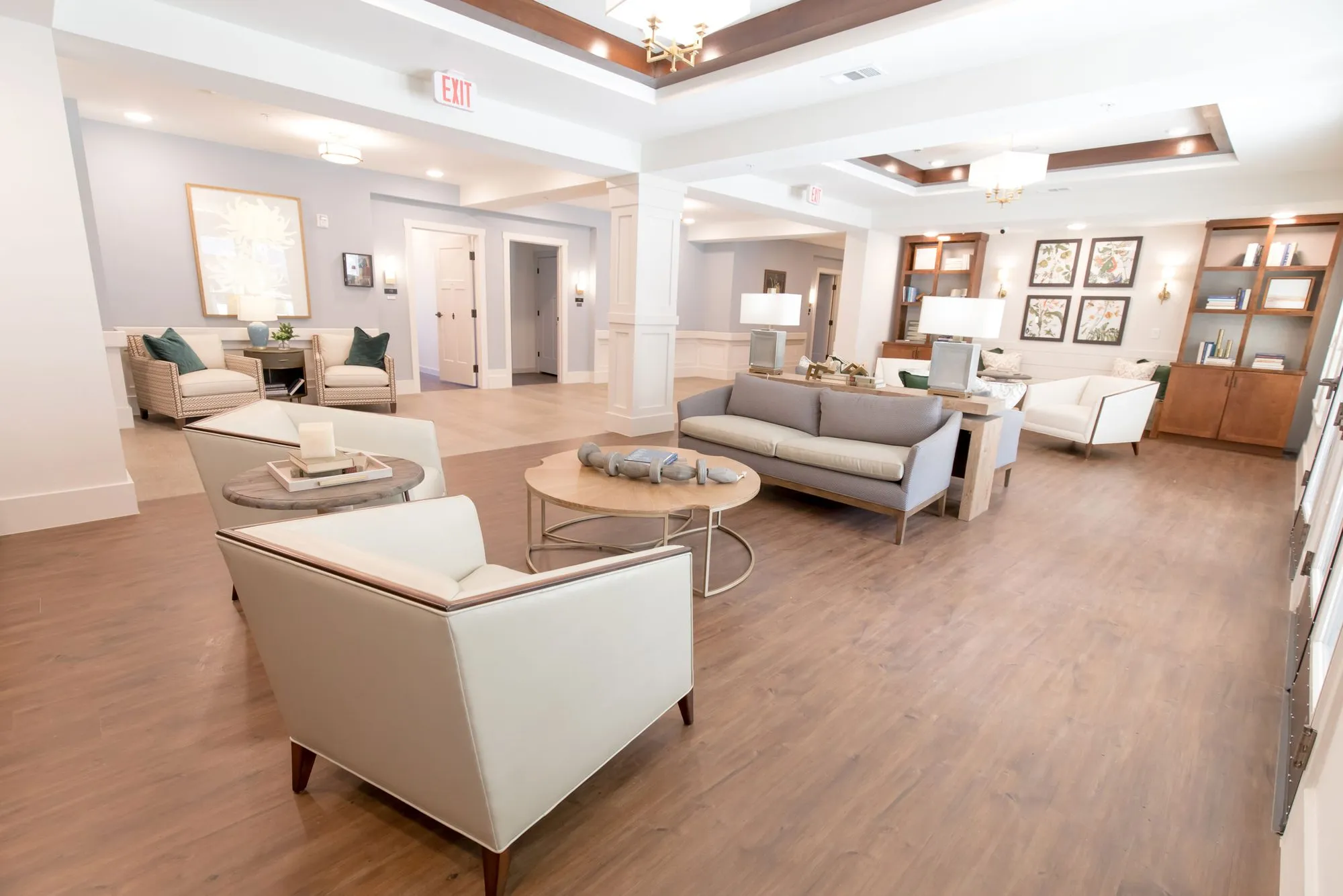 Senior living living room with white armchairs, a grey sofa and round tables under tray ceilings, wood bookcases beyond