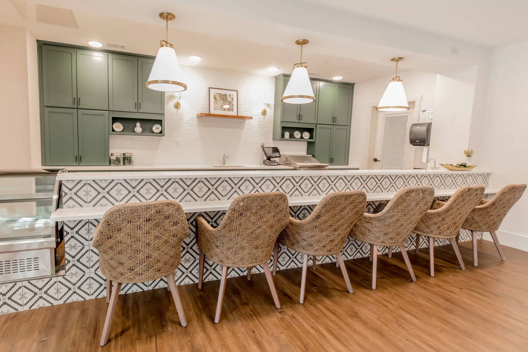 Senior living bistro with woven counter chairs along a patterned tile bar front below sage cabinetry and glass pendants