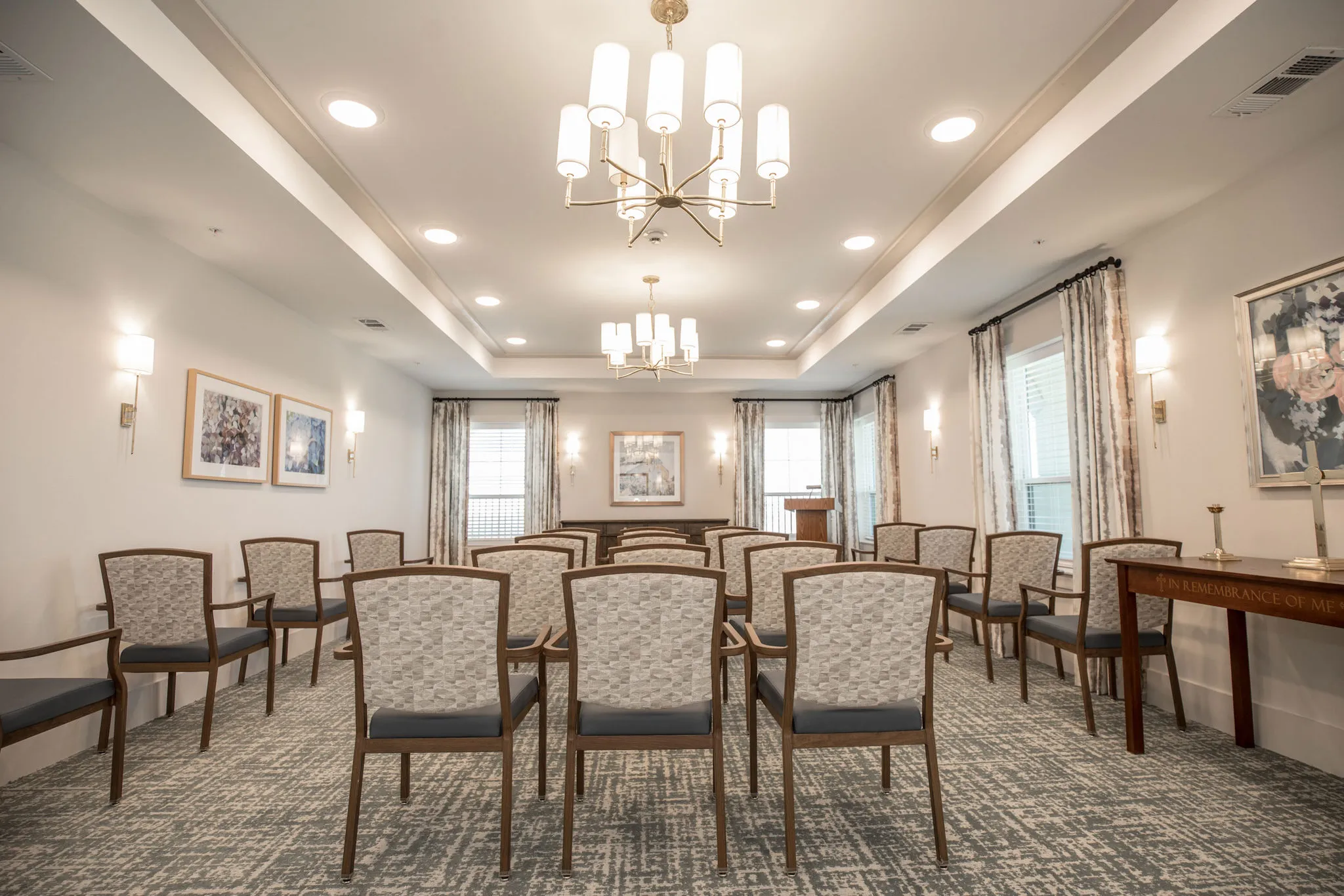 Senior living chapel with rows of wood chairs under twin chandeliers between draped windows