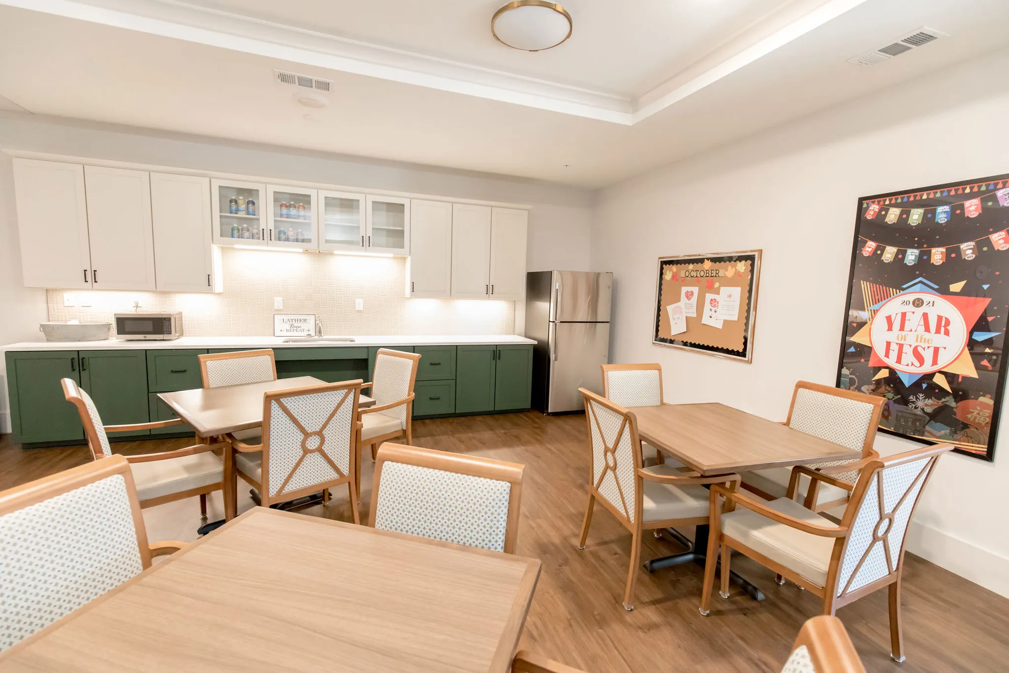 Senior living kitchenette with sage lower cabinets and a fridge behind two light oak tables, a bulletin board at right