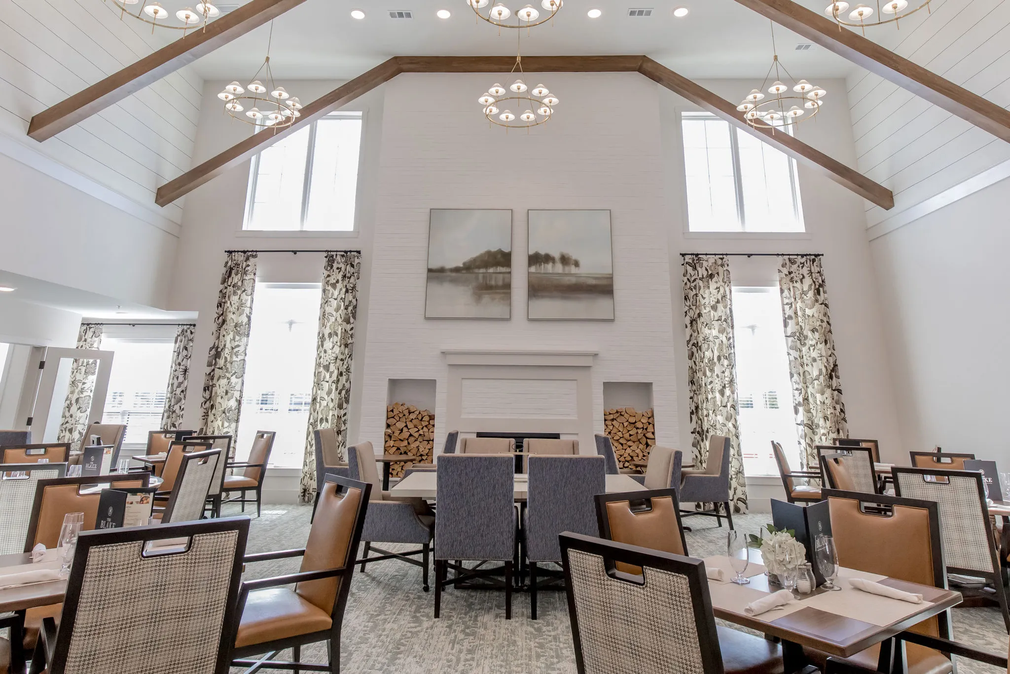 Senior living dining room with a tall shiplap ceiling, a white brick fireplace between stacked firewood and brass chandeliers