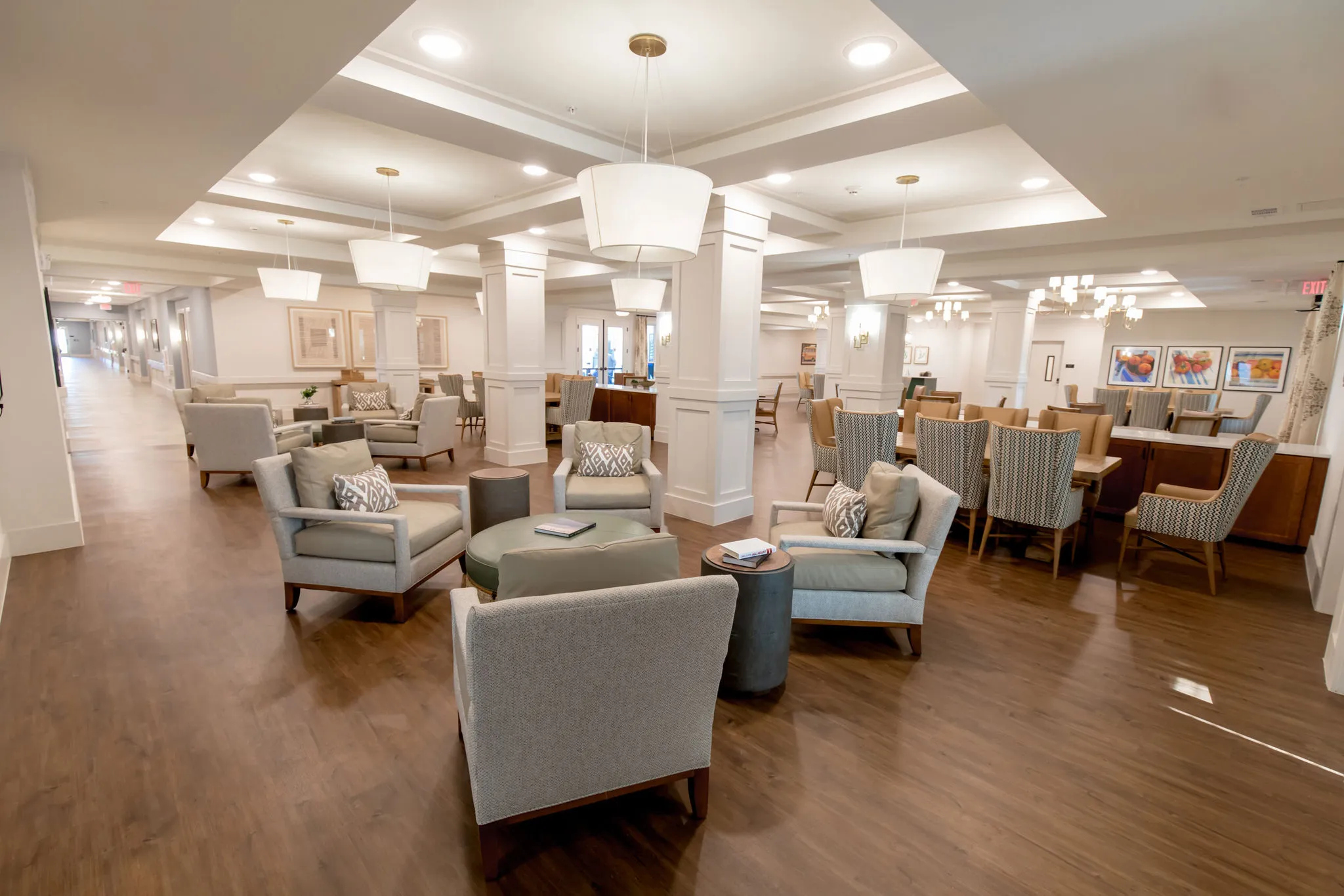 Senior living parlor with grey lounge chairs and drum pendants along the open living and dining wing