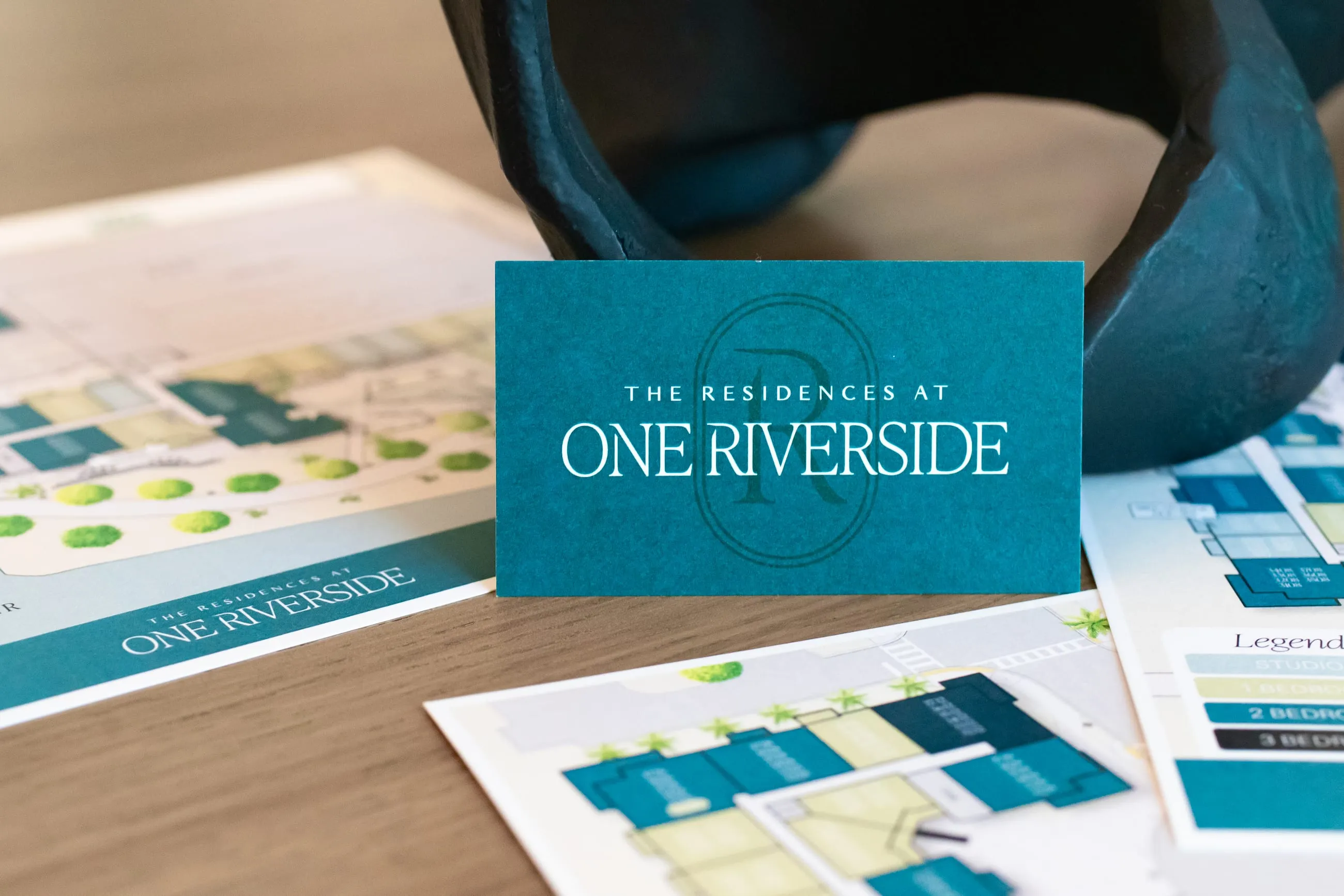 A teal business card propped against a dark sculptural vase on a wood desk, over printed site plans and brochures in matching teal and green