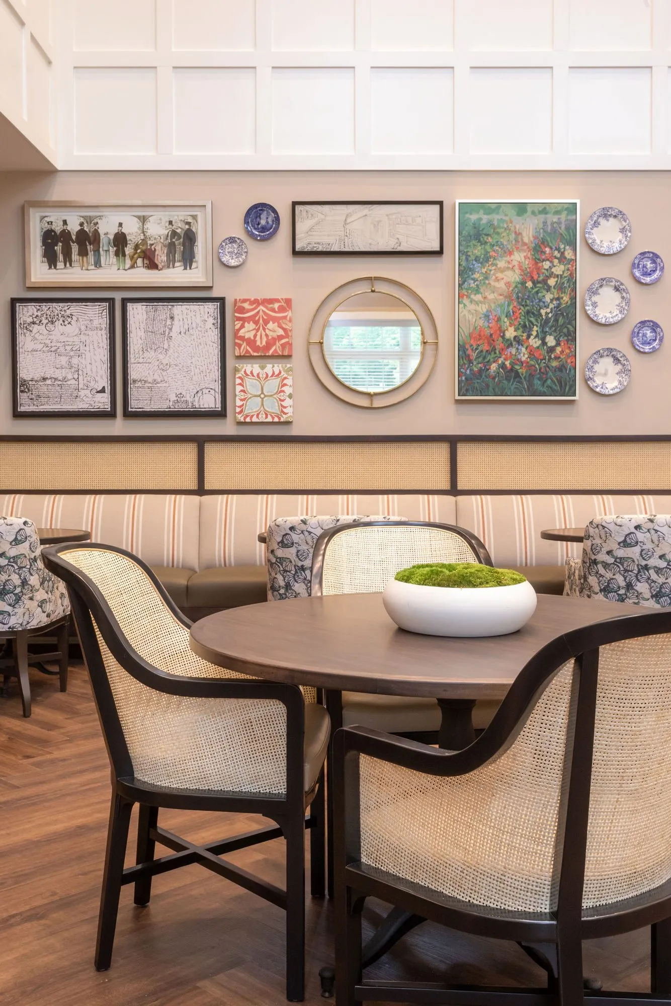 Senior living dining room with cane-back chairs at a round table below a gallery of blue and white plates and a round mirror