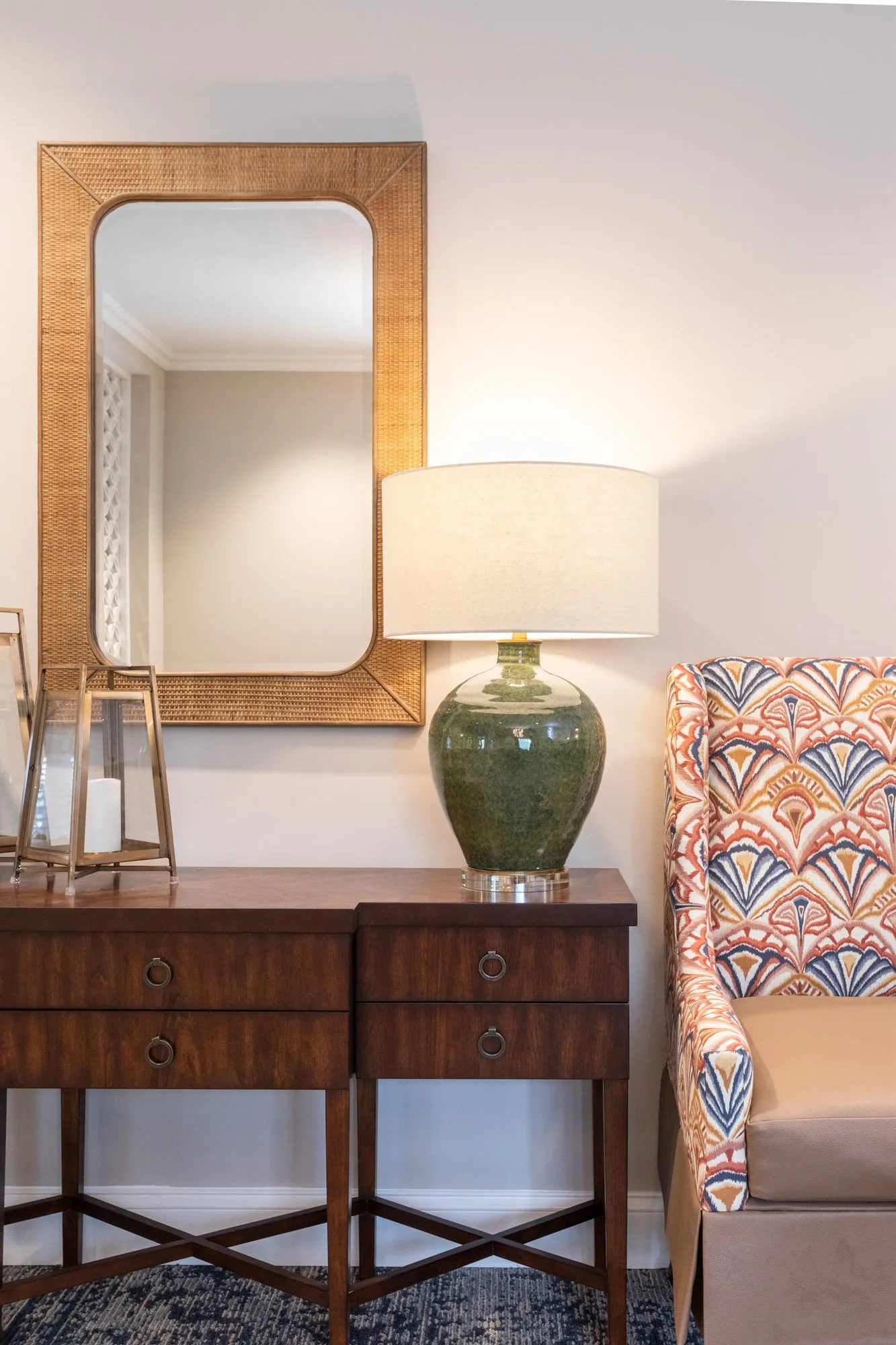 Senior living interior detail: wood console with a green ceramic lamp beneath a rattan-framed mirror by a patterned armchair