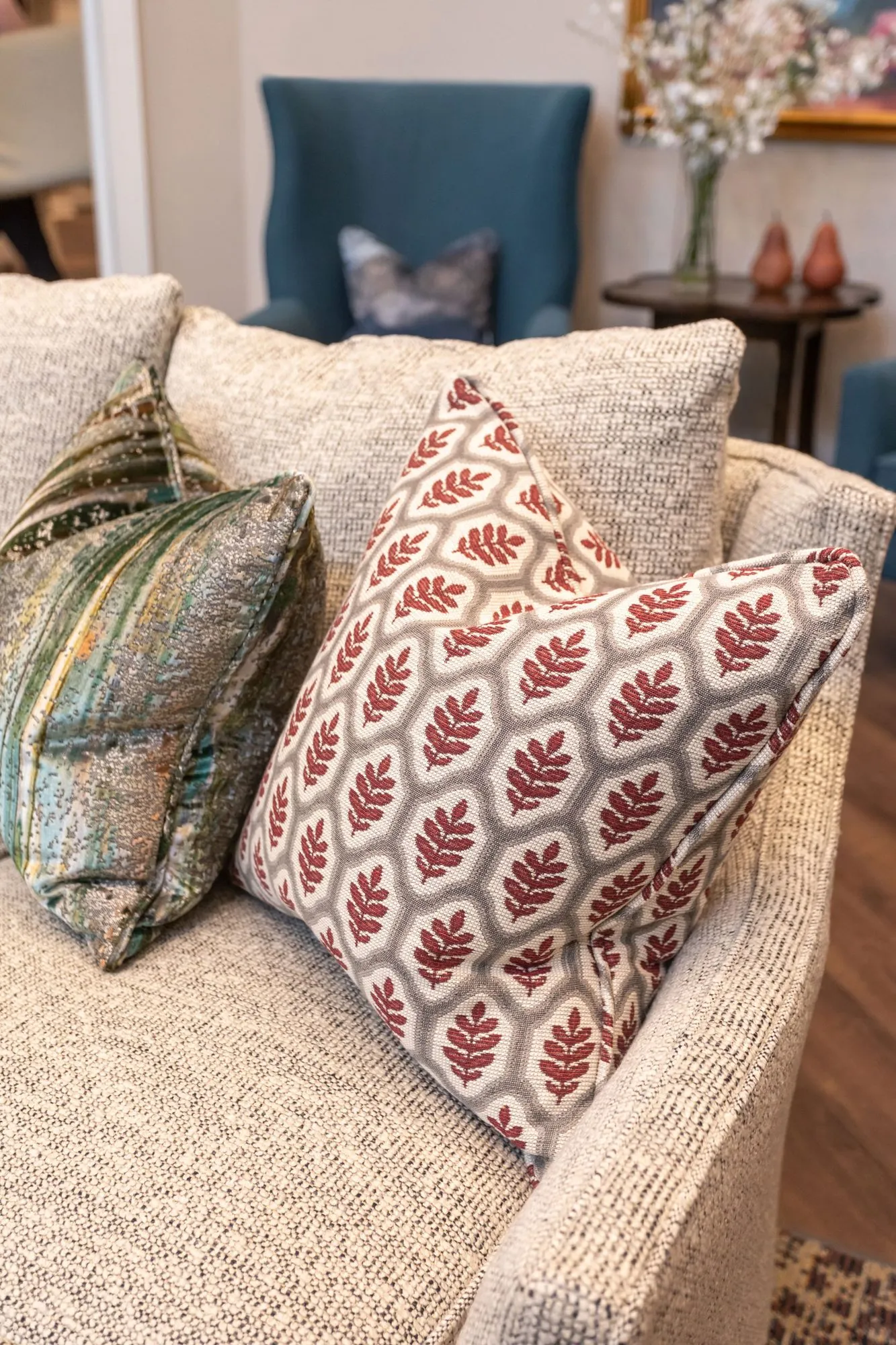 Senior living interior detail: cream boucle sofa with a red leaf-print pillow and a teal velvet pillow, a blue wing chair