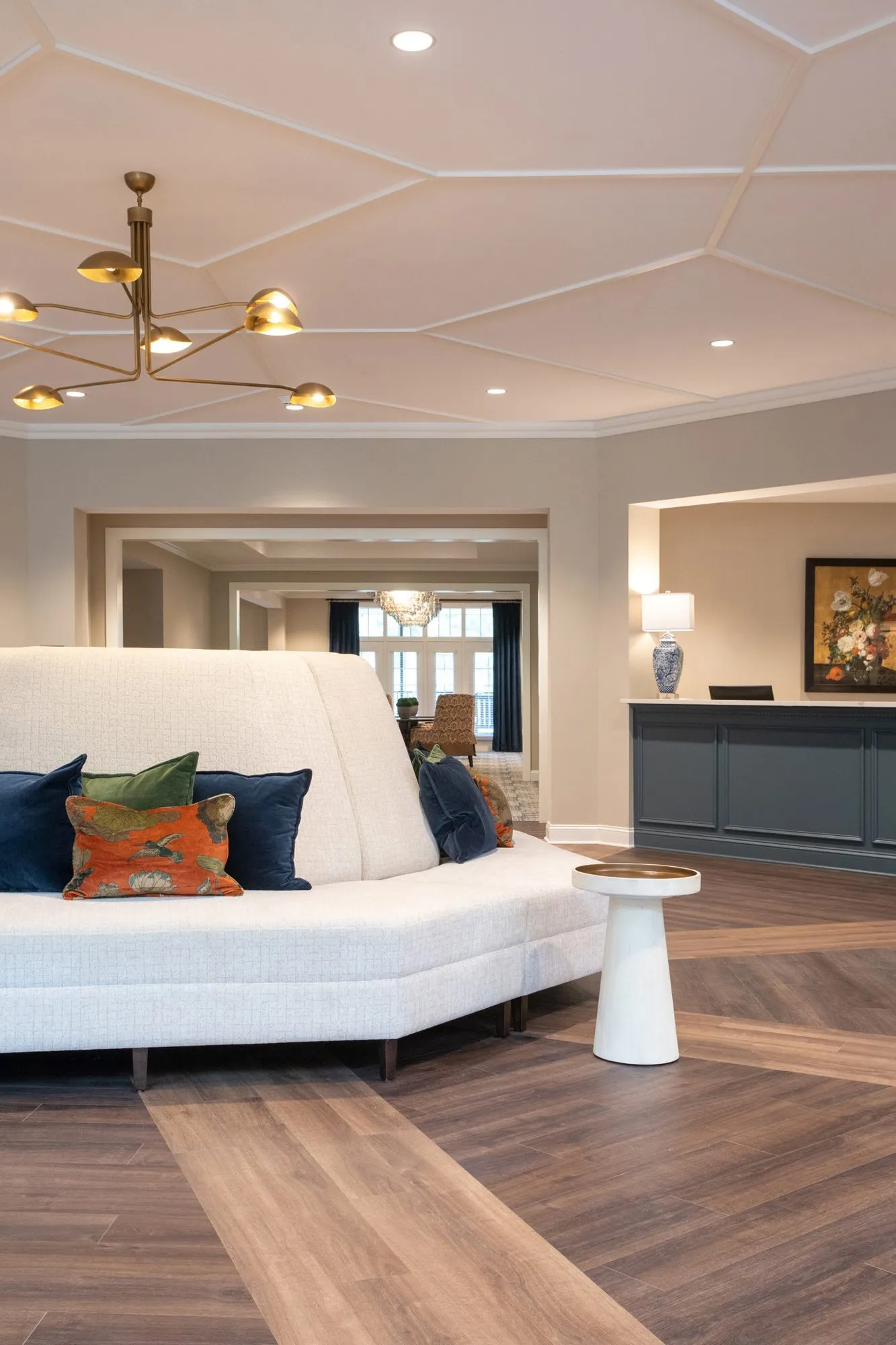 Senior living lobby with a curved white settee and navy and orange pillows on a herringbone floor, reception counter beyond