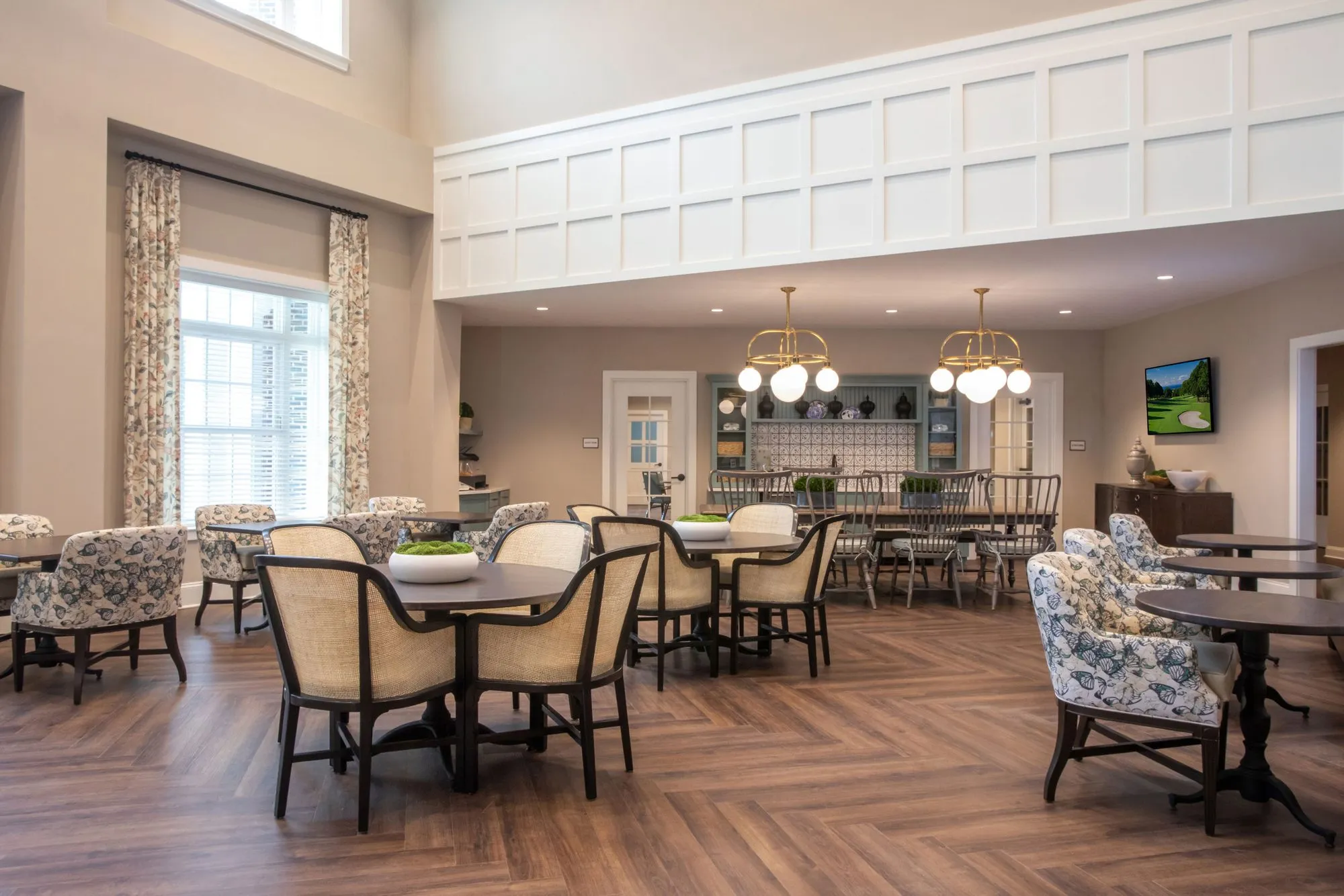 Senior living dining area under white panelling and brass globe chandeliers, cane-back and floral chairs at round tables