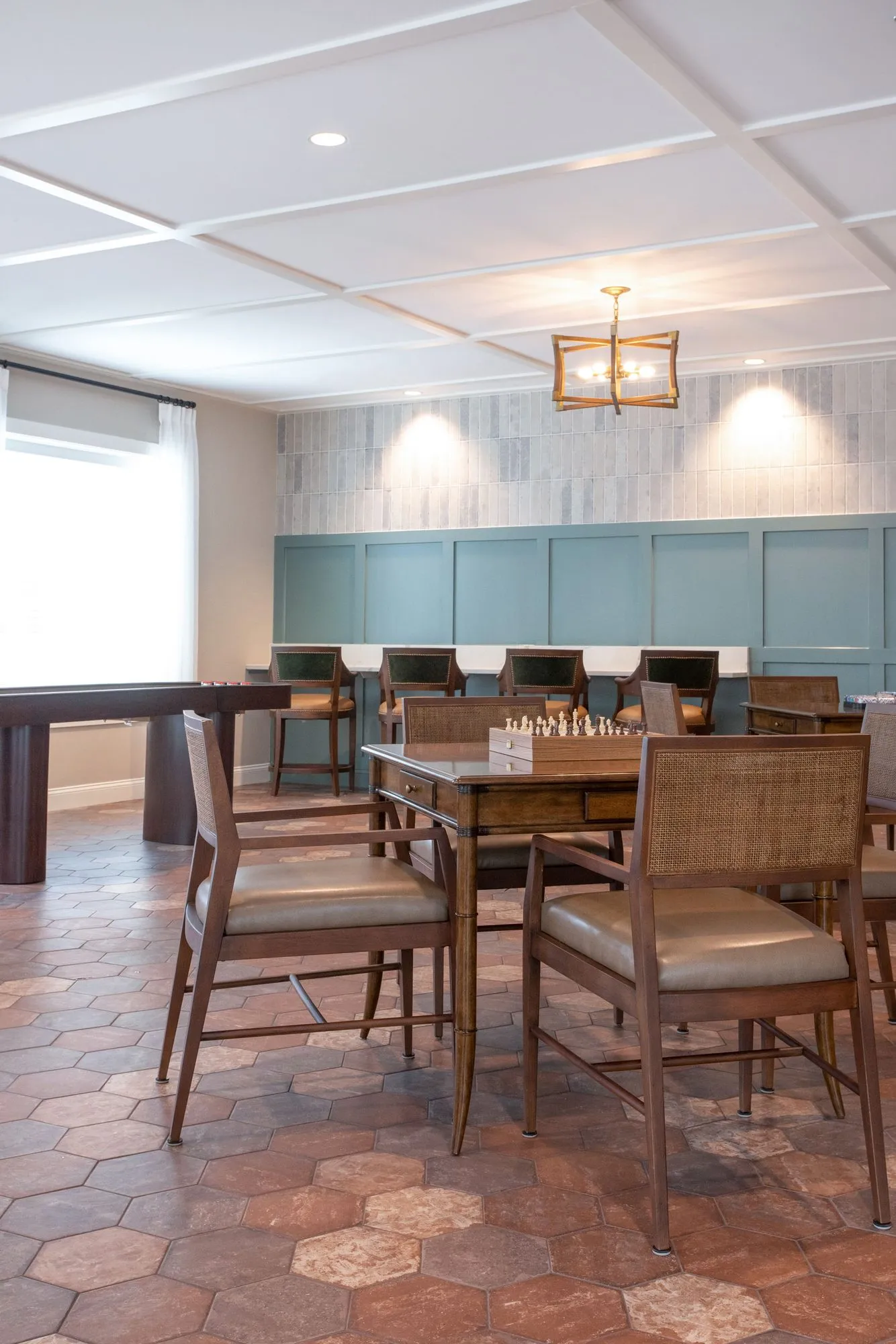 Senior living game room on terracotta hexagon tile with a chess table and cane-back chairs, a shuffleboard table by the wall