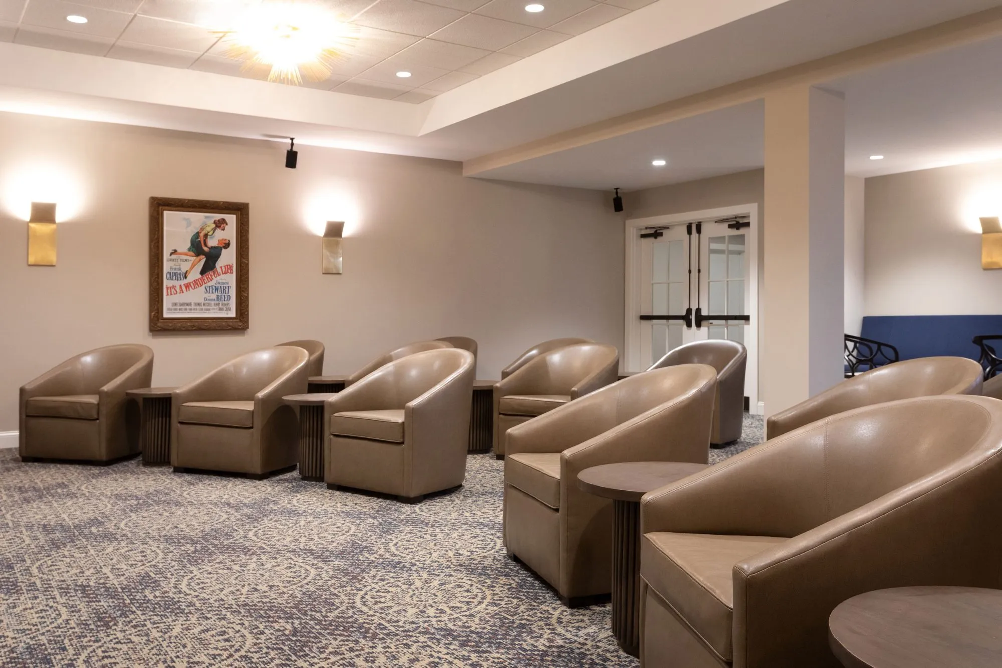 Senior living theater with rows of taupe leather barrel chairs and side tables on patterned carpet, a framed film poster