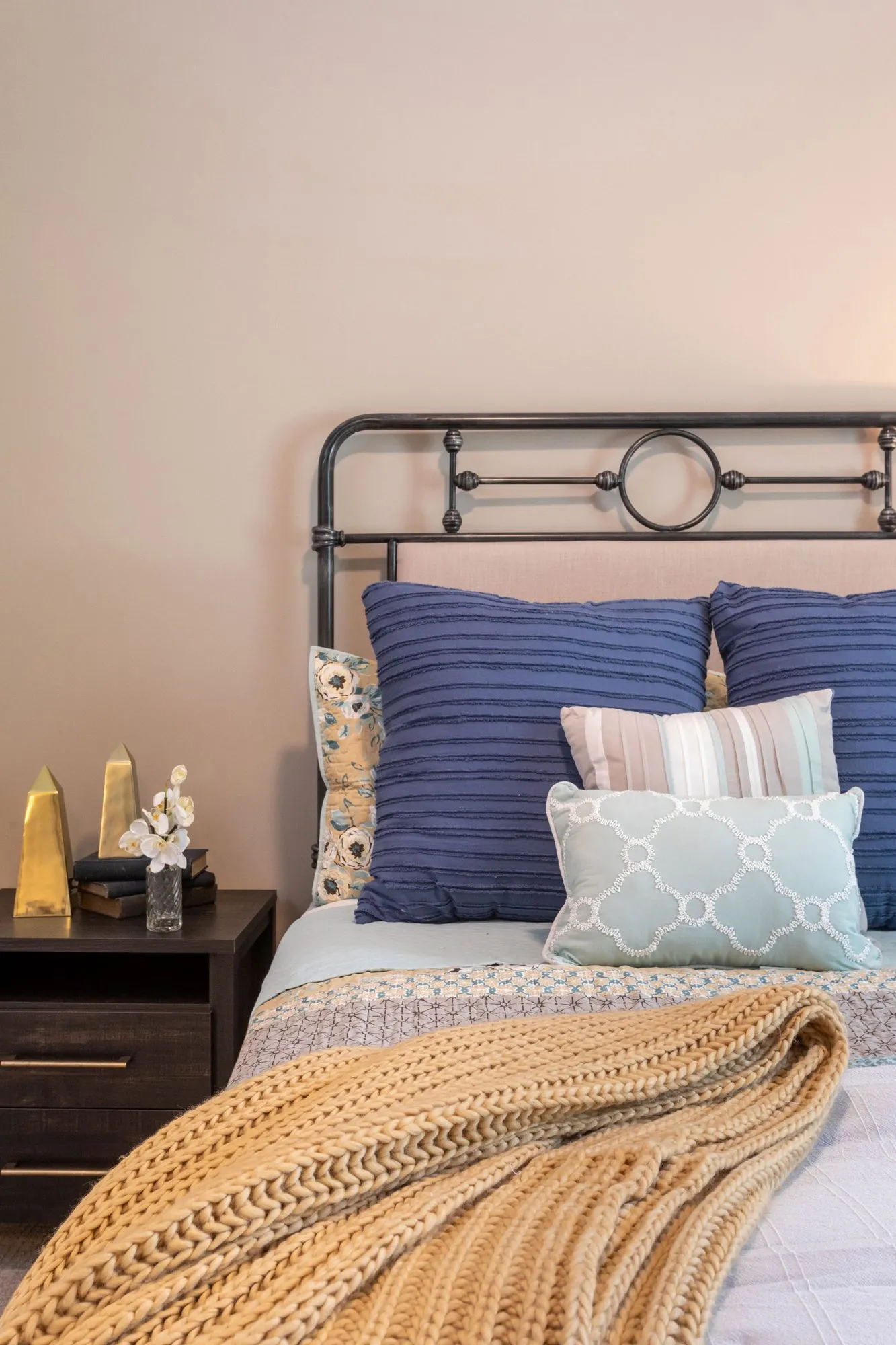 Senior living bedroom with an iron bed, blue and pale aqua pillows and a mustard knit throw beside a dark nightstand